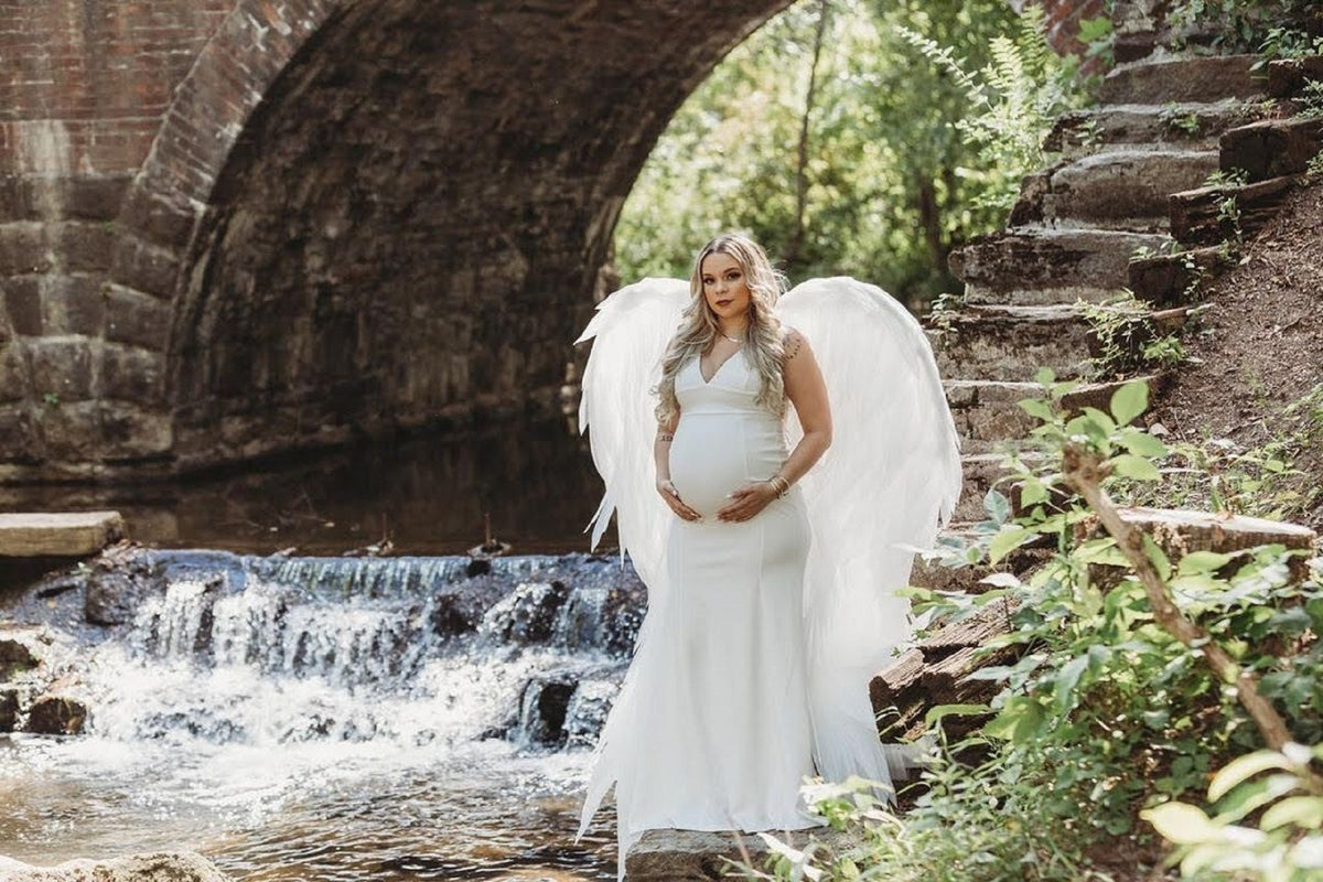 May include: A pregnant woman wearing a white dress and angel wings stands in front of a waterfall and stone bridge.