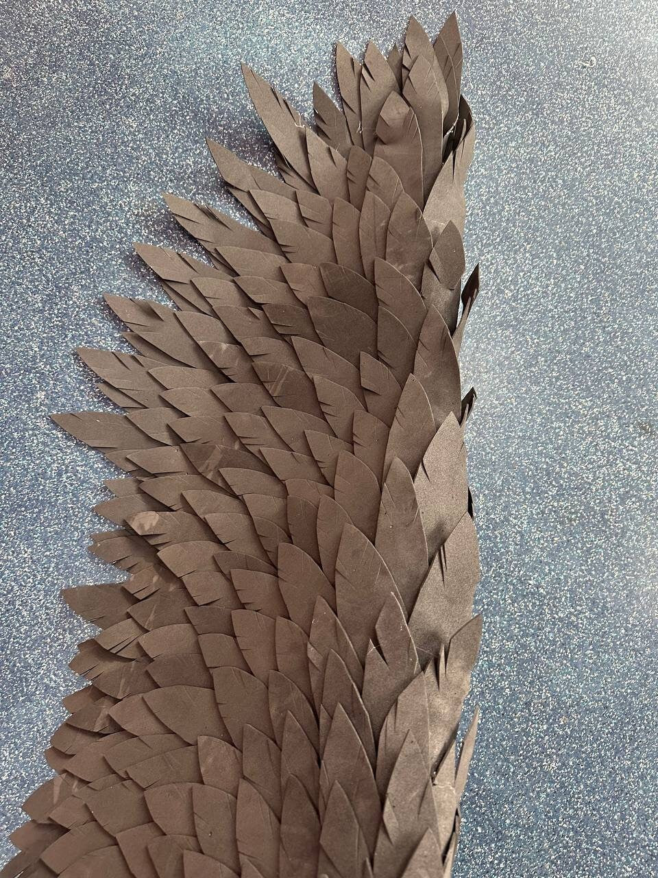 May include: A close-up of a brown paper wing with a layered, feathered design. The wing is made of many individual paper feathers that are layered on top of each other to create a realistic look.