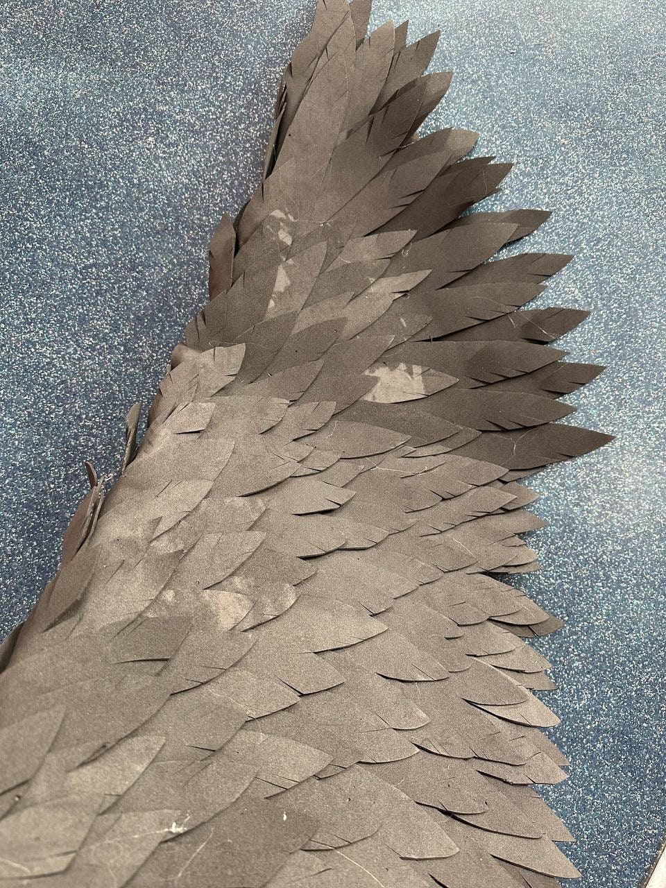 May include: A close-up of a black paper wing with a layered, feathered design. The wing is made of multiple pieces of paper that are layered on top of each other to create a 3D effect.
