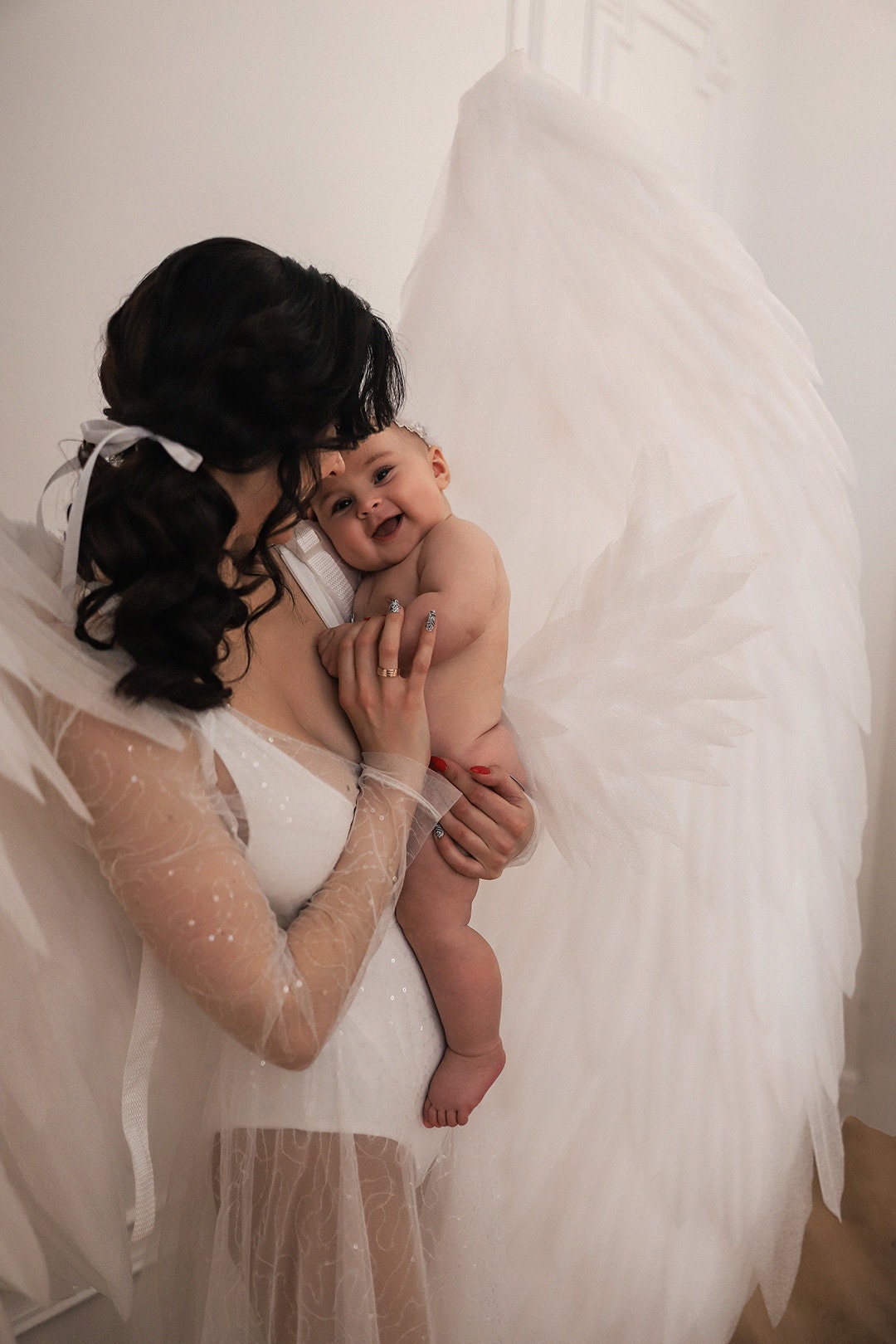 May include: A woman in a white, sparkly, sheer dress holds a baby in her arms. The baby is smiling and looking at the camera. The woman is wearing a white headband and has long, dark hair. The background is a white wall.