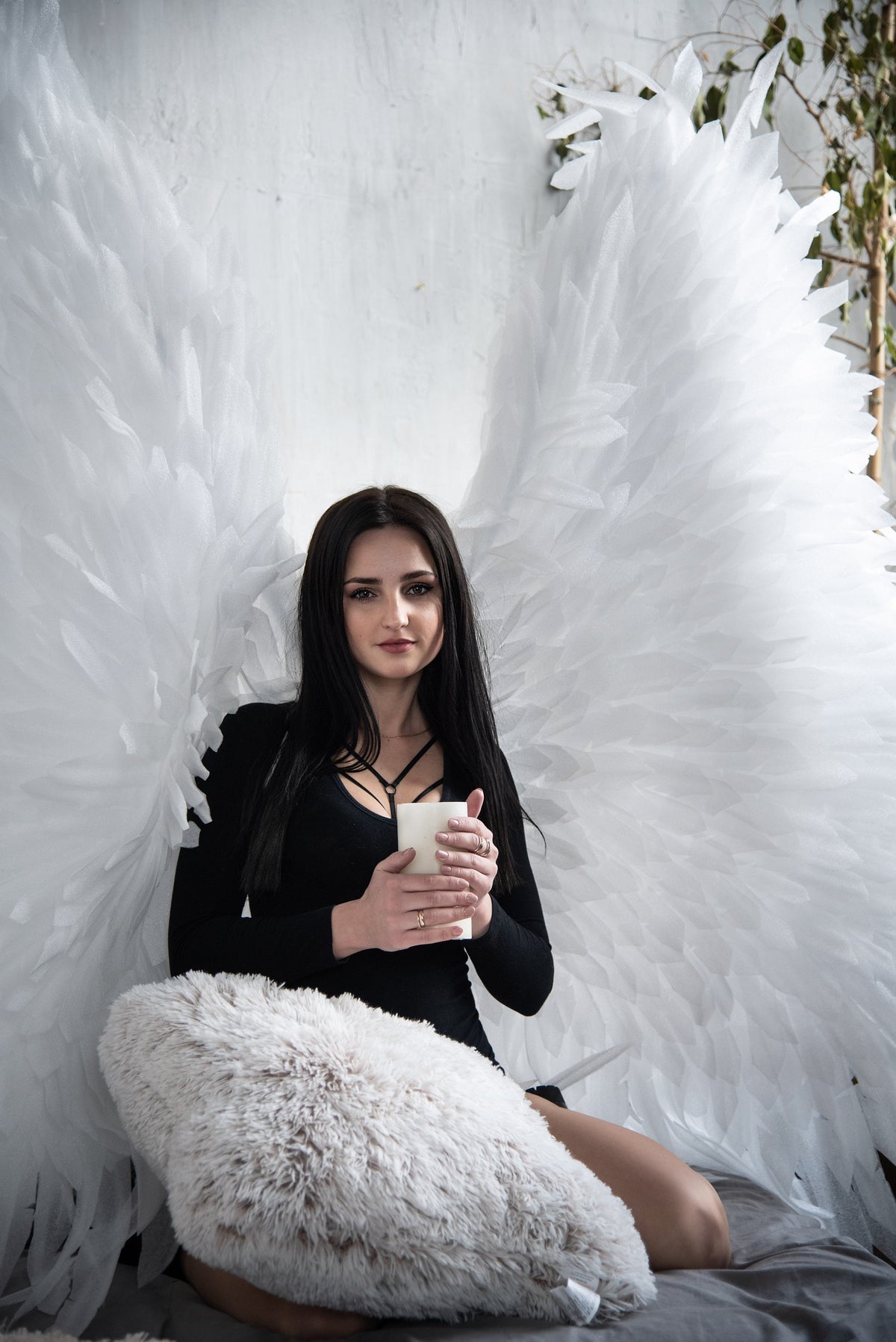 May include: A woman with long black hair is sitting on a white fluffy pillow with large white angel wings behind her. She is holding a white candle in her hands.