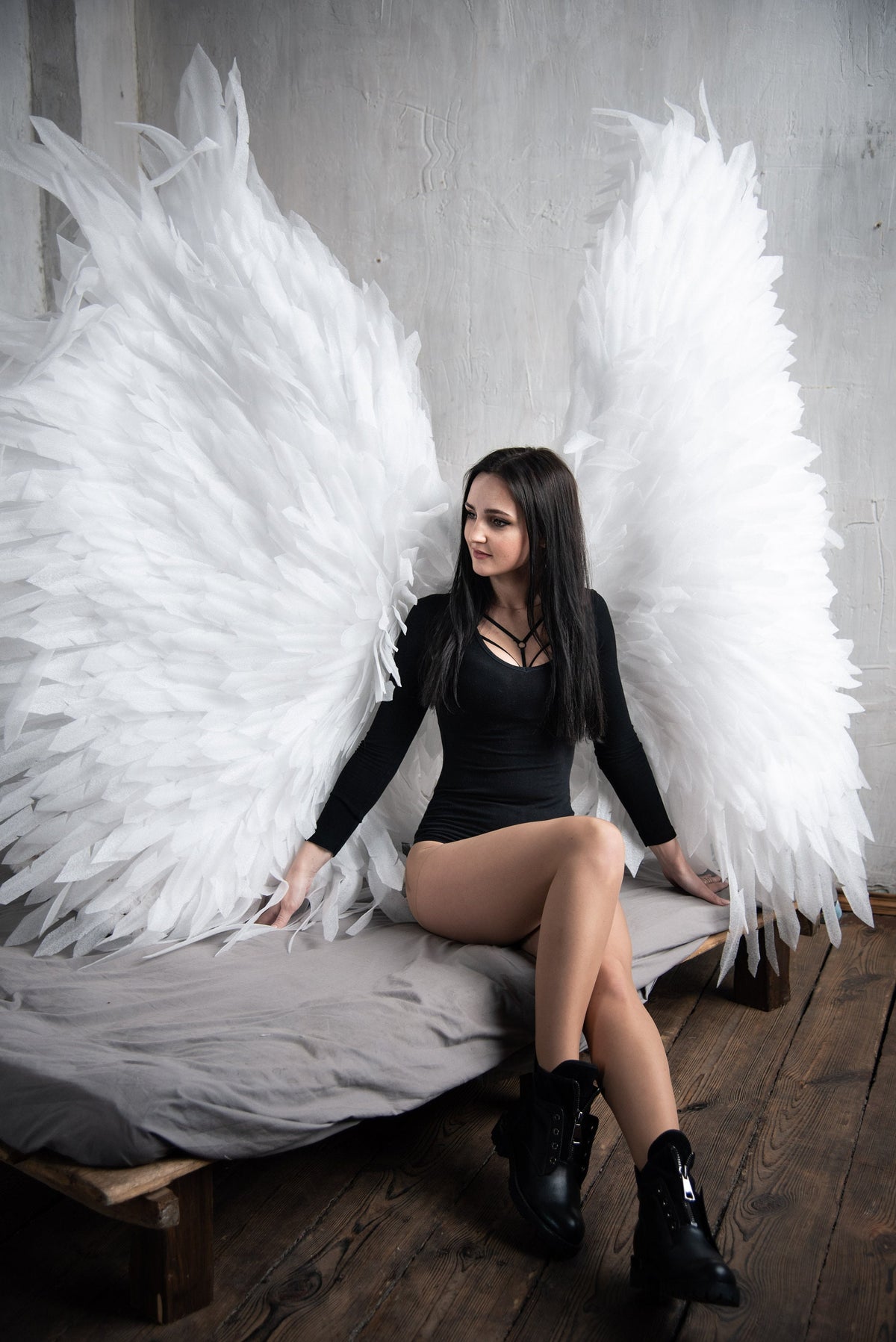 May include: A woman wearing a black bodysuit and black boots sits on a bed with large white angel wings behind her.