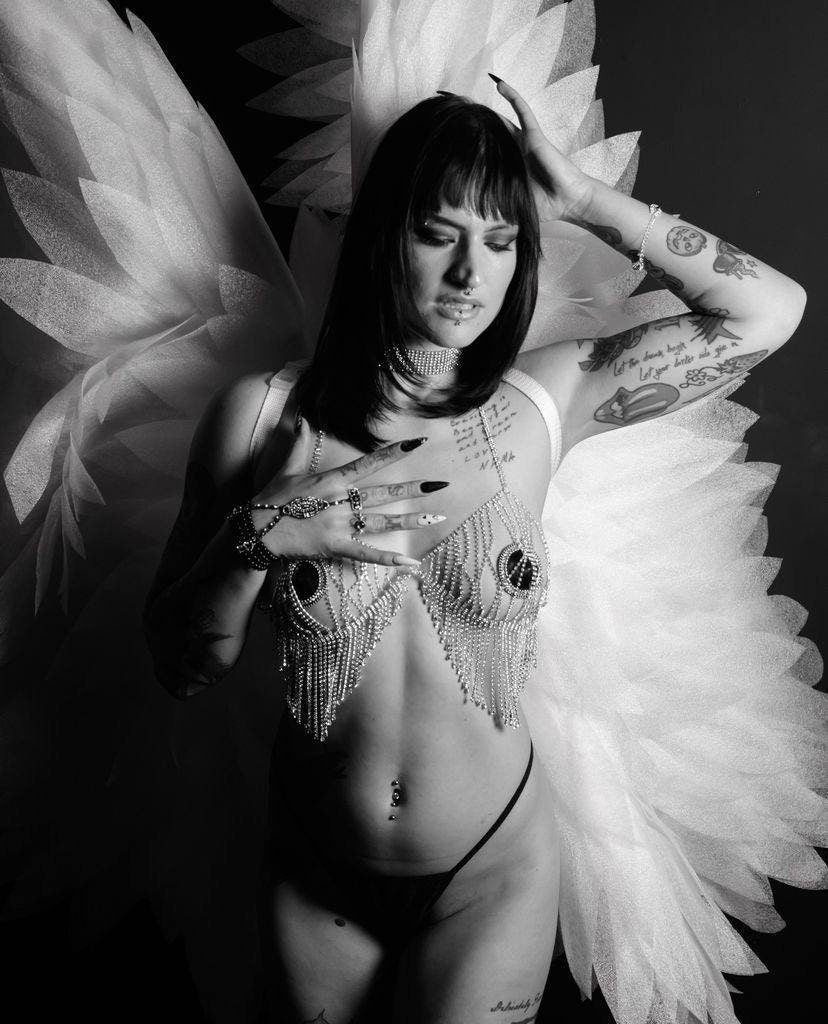 May include: Black and white image of a woman wearing a jeweled bra with dangling rhinestones and a matching choker. She has long black hair, dark nail polish, and a large white feathered wing. The image is a studio shot.