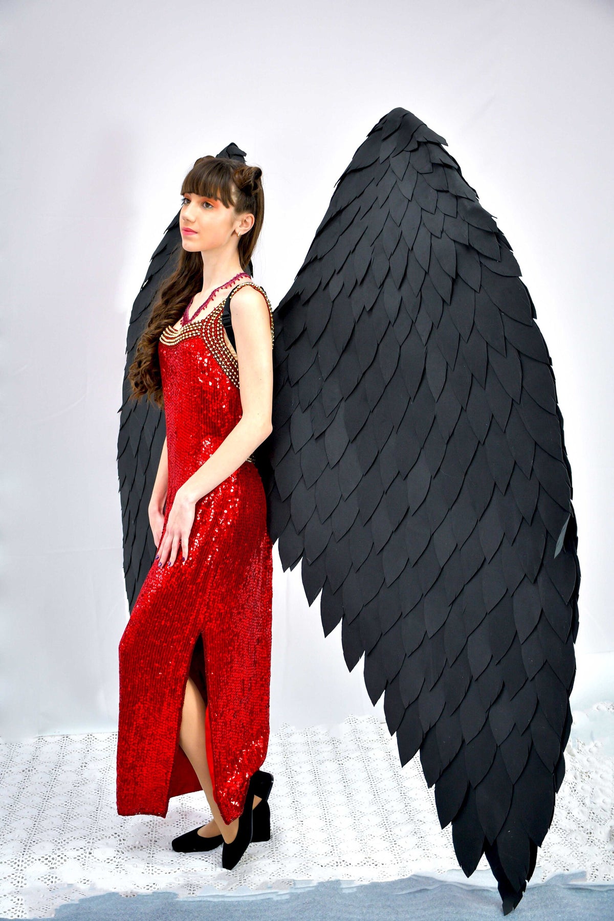 May include: A woman wearing a red sequined dress with a black feather angel wing costume. The wings are large and detailed, with many individual feathers.