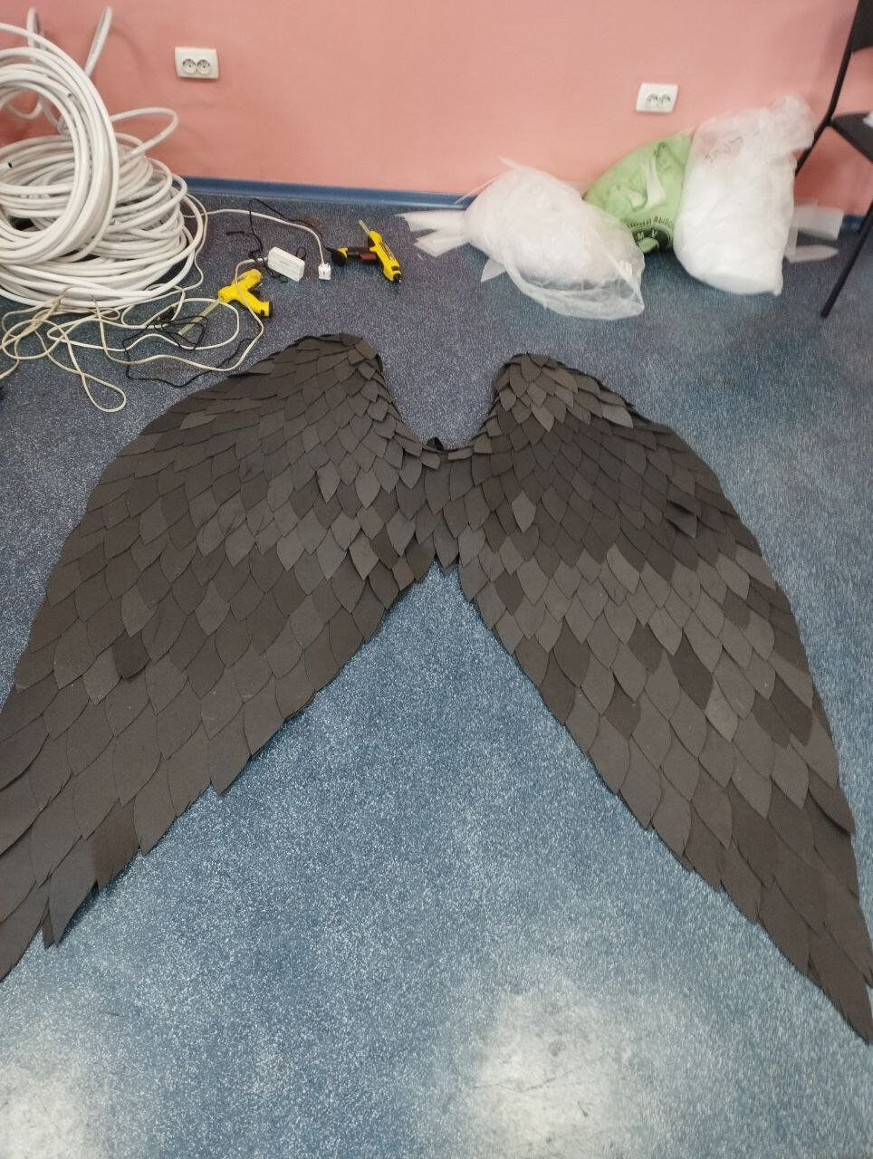 May include: A pair of large, black, paper wings with a scaly texture. The wings are laid out flat on a blue floor.