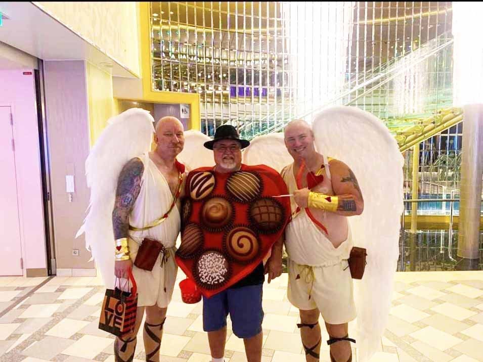 May include: Three people dressed in costumes for a party. Two people are wearing white togas with angel wings. One person is wearing a red heart-shaped costume with chocolate candies on it. The person in the middle is holding a red cup.