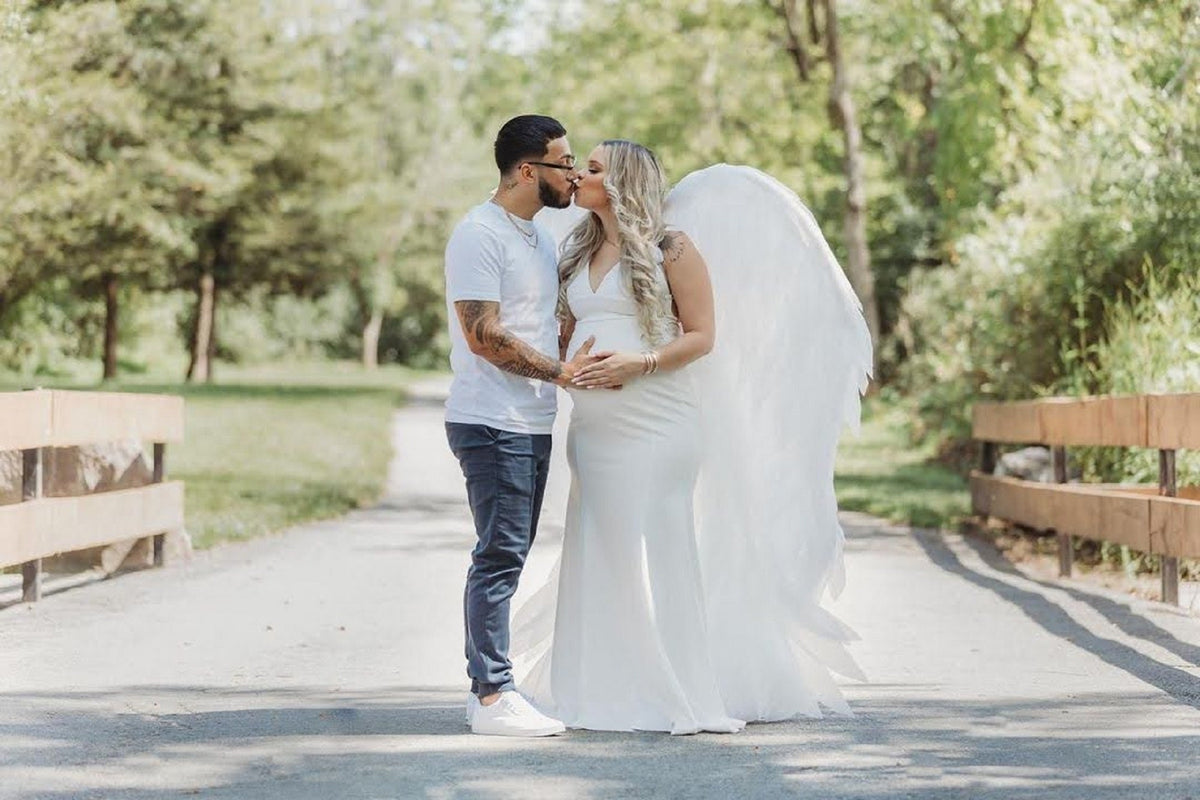 May include: A pregnant woman in a white dress with angel wings kisses her partner. They are standing on a path in a park.