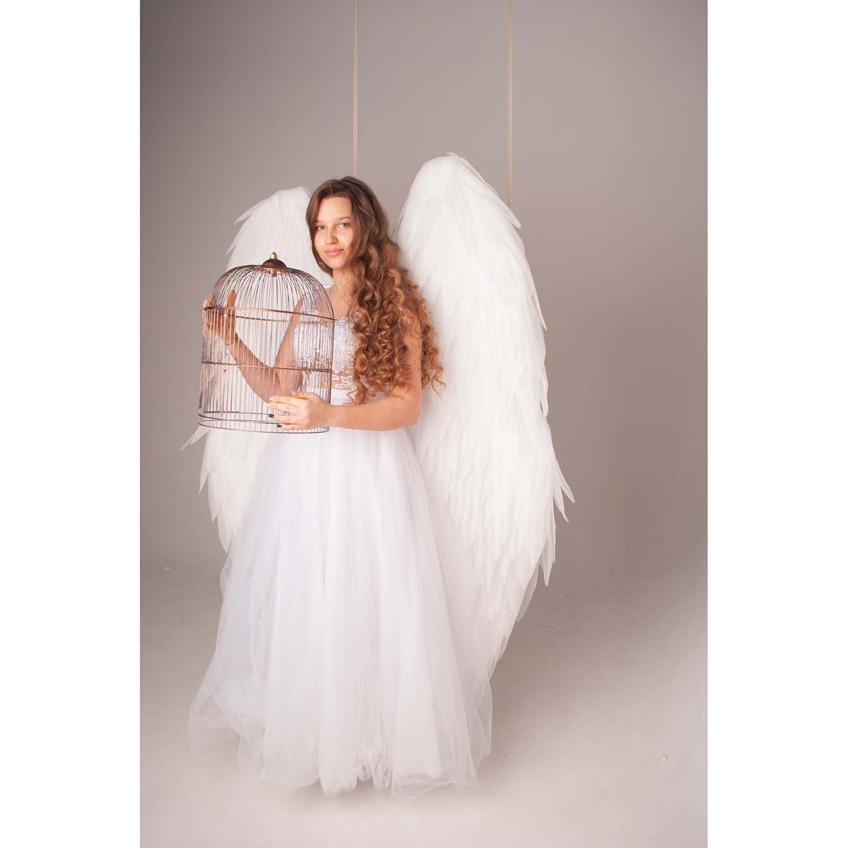 May include: A young woman wearing a white dress and large white angel wings holds a birdcage in front of her. The cage is made of metal and has a rounded top.