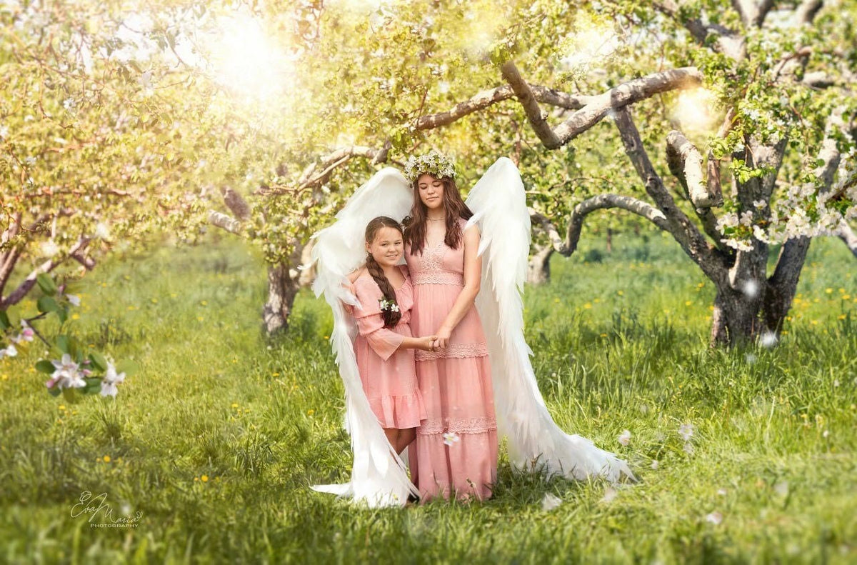 May include: Two girls wearing pink dresses and white angel wings stand in a field of green grass with a blooming tree behind them.