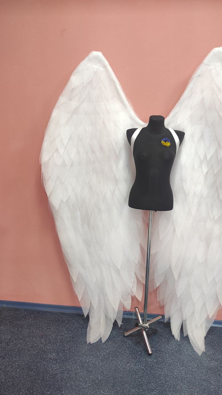 May include: A pair of large white angel wings with a black mannequin wearing a white strap in the center. The mannequin is wearing a blue and yellow pin.