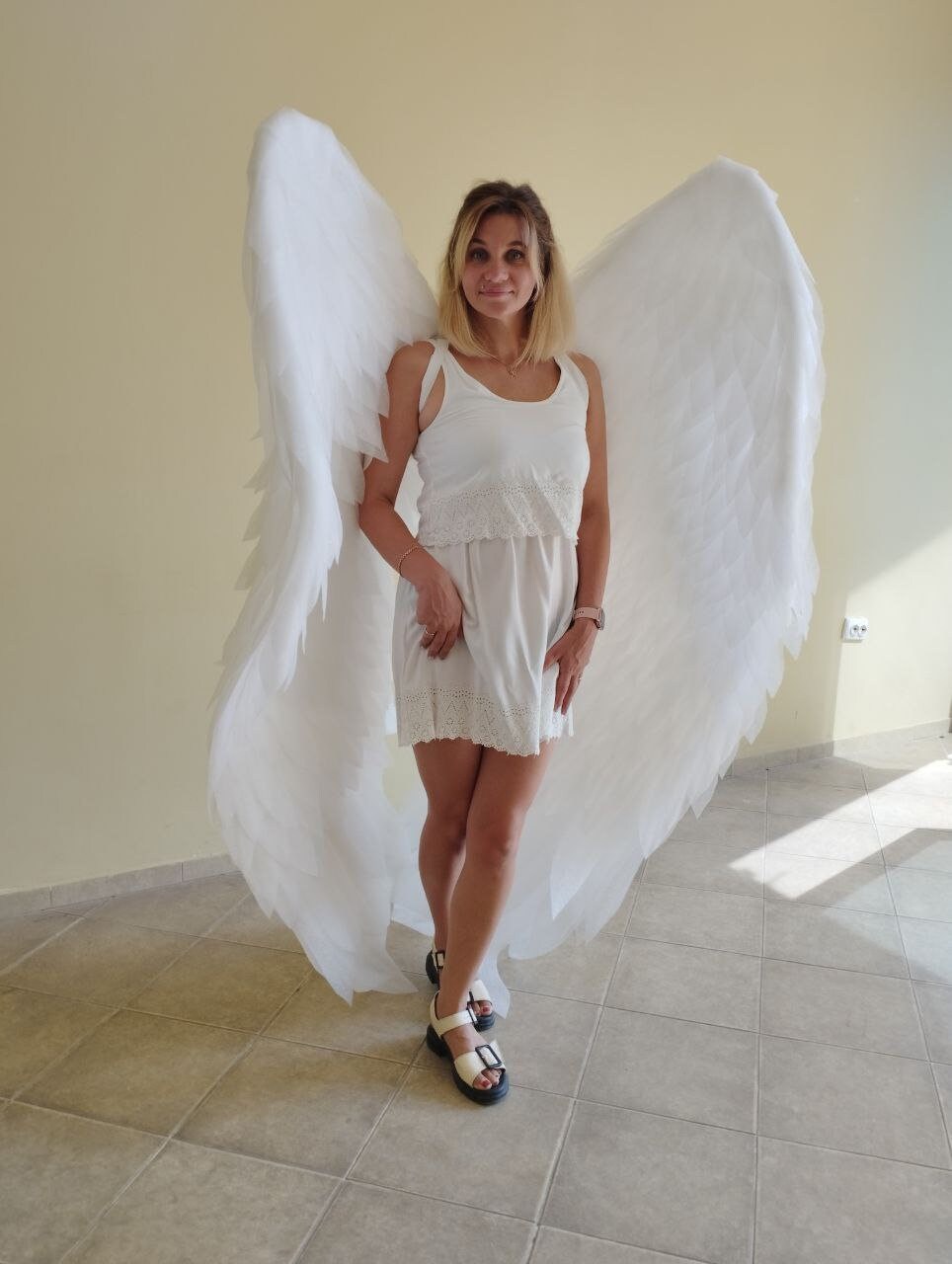 May include: A woman wearing a white dress and white angel wings. The wings are large and feathery, and the woman is standing on a tiled floor.