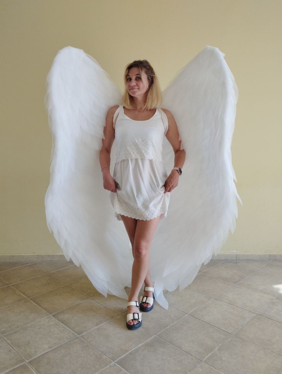 May include: A woman wearing a white dress and white sandals stands in front of a light yellow wall. She is wearing large white angel wings.