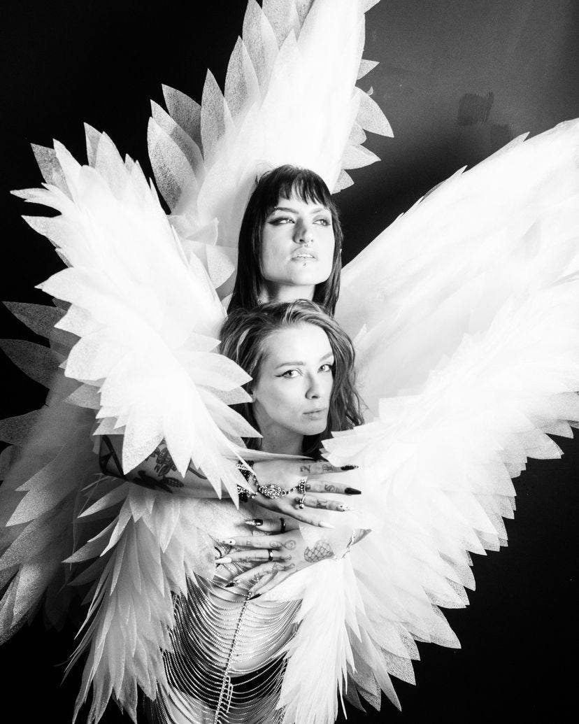 May include: Black and white image of two people embracing, wearing large, feathery white angel wings. The wings are layered and textured. The people are in close proximity, with one person's arms wrapped around the other. The background is solid black.