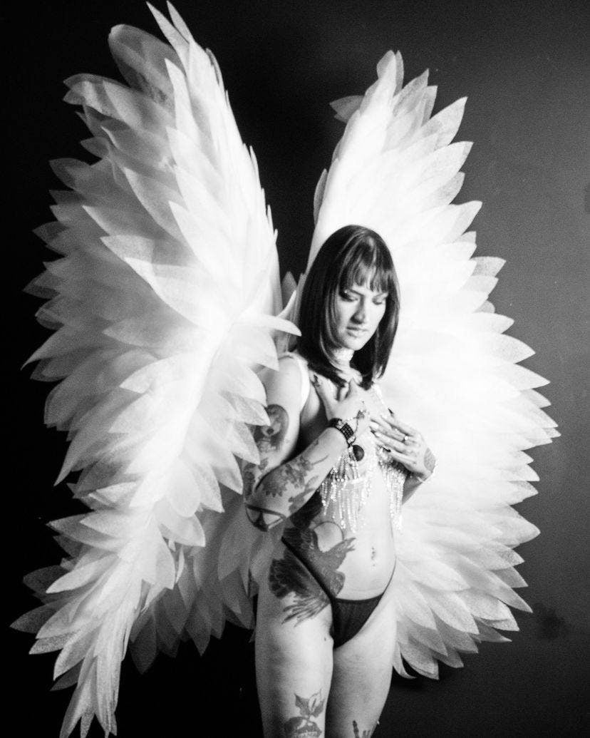 May include: A black and white image of large, white, feathery angel wings. The wings are the focal point, with a person wearing them. The person is wearing a black thong and has tattoos on their body.
