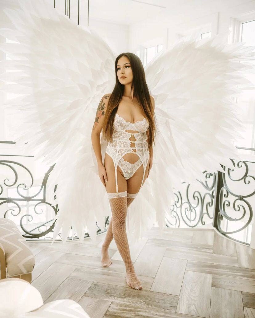 May include: A woman wearing a white lace lingerie set with garter straps and fishnet stockings, posing with large white feathered angel wings. The background features a light-colored interior with a decorative railing.