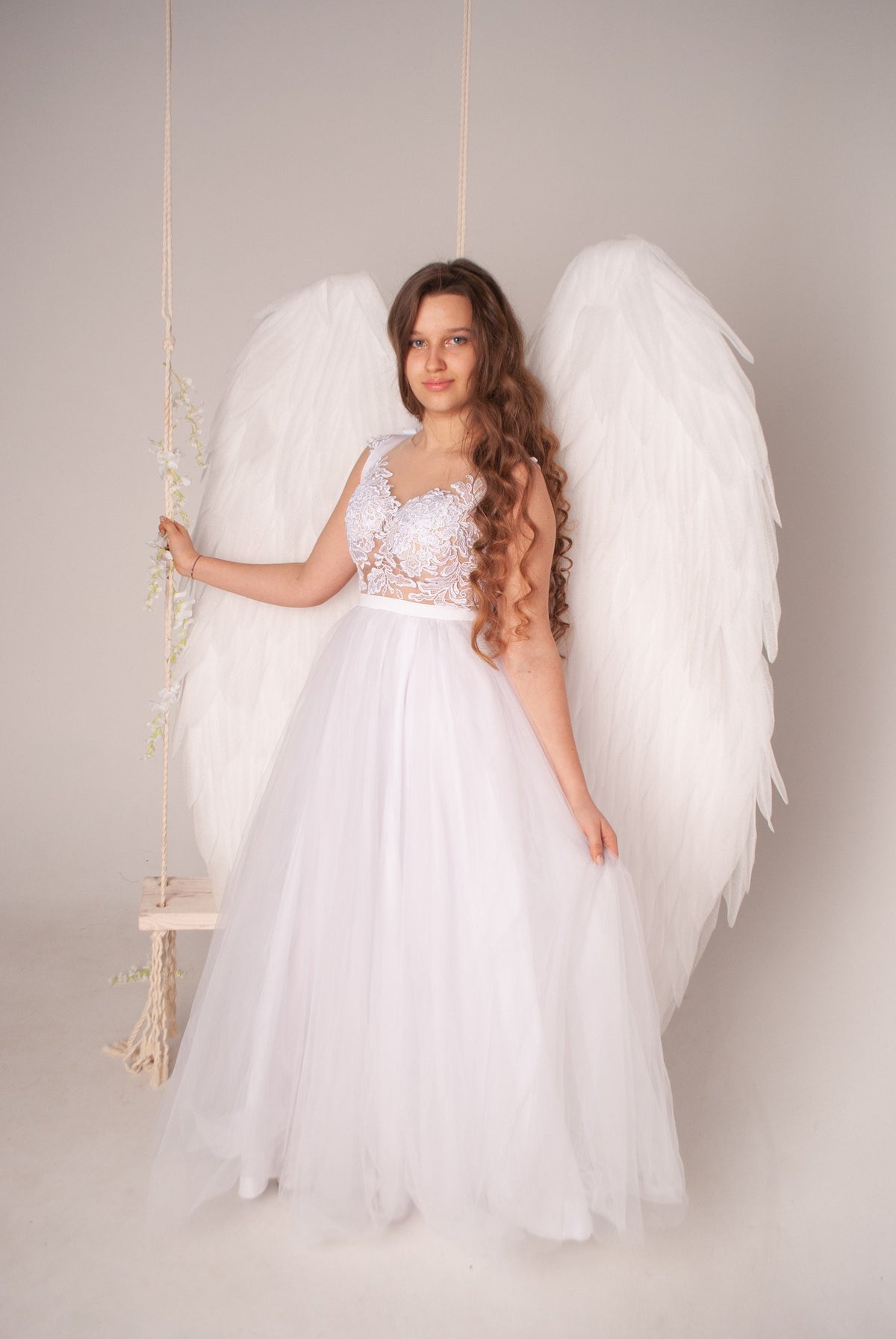 May include: A young woman wearing a white lace dress and large white angel wings. She is standing in front of a white background.