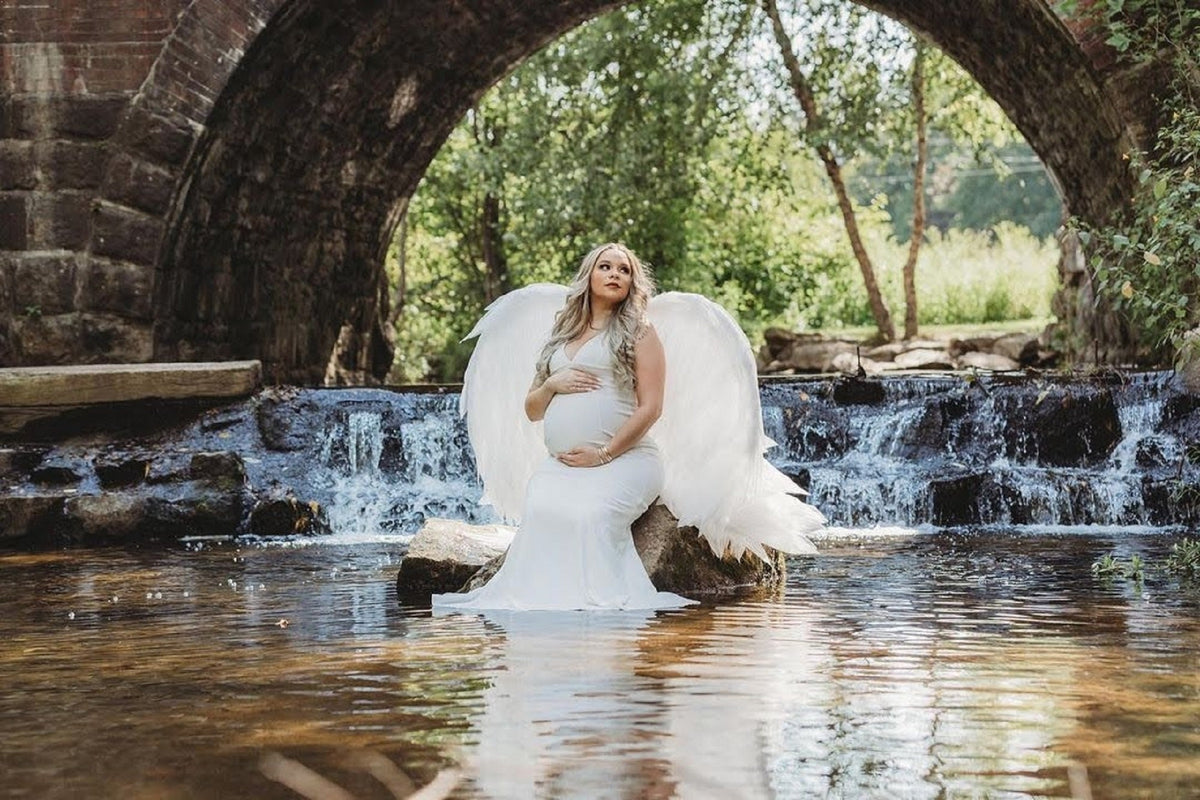 May include: A pregnant woman wearing a white dress and angel wings sits on a rock in a stream. The water is flowing over a small waterfall behind her.