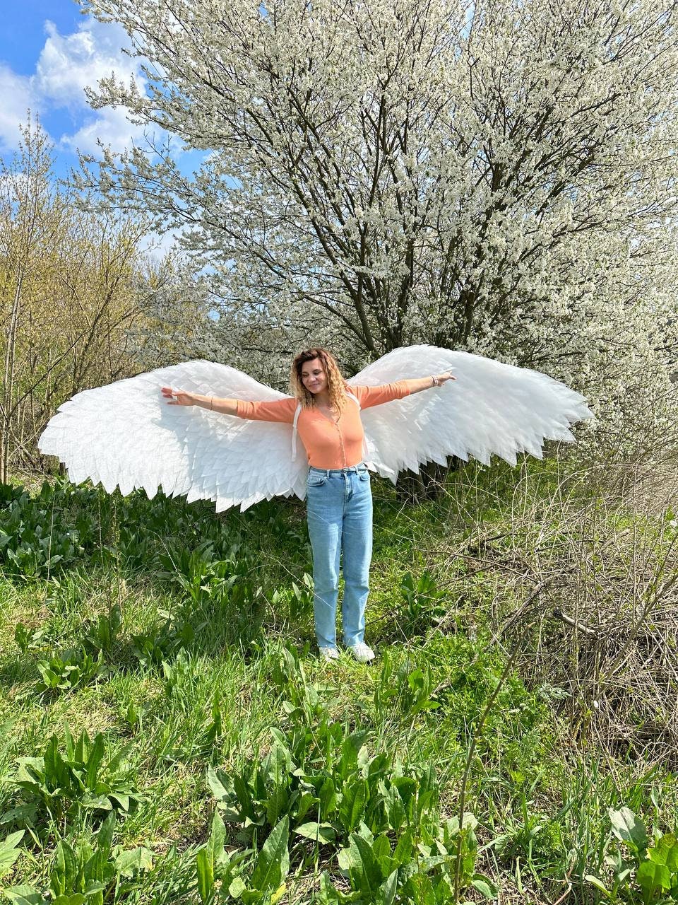 May include: A person wearing a pink shirt and blue jeans is standing in a grassy field with a white tree in the background. They are holding large white wings out to their sides.