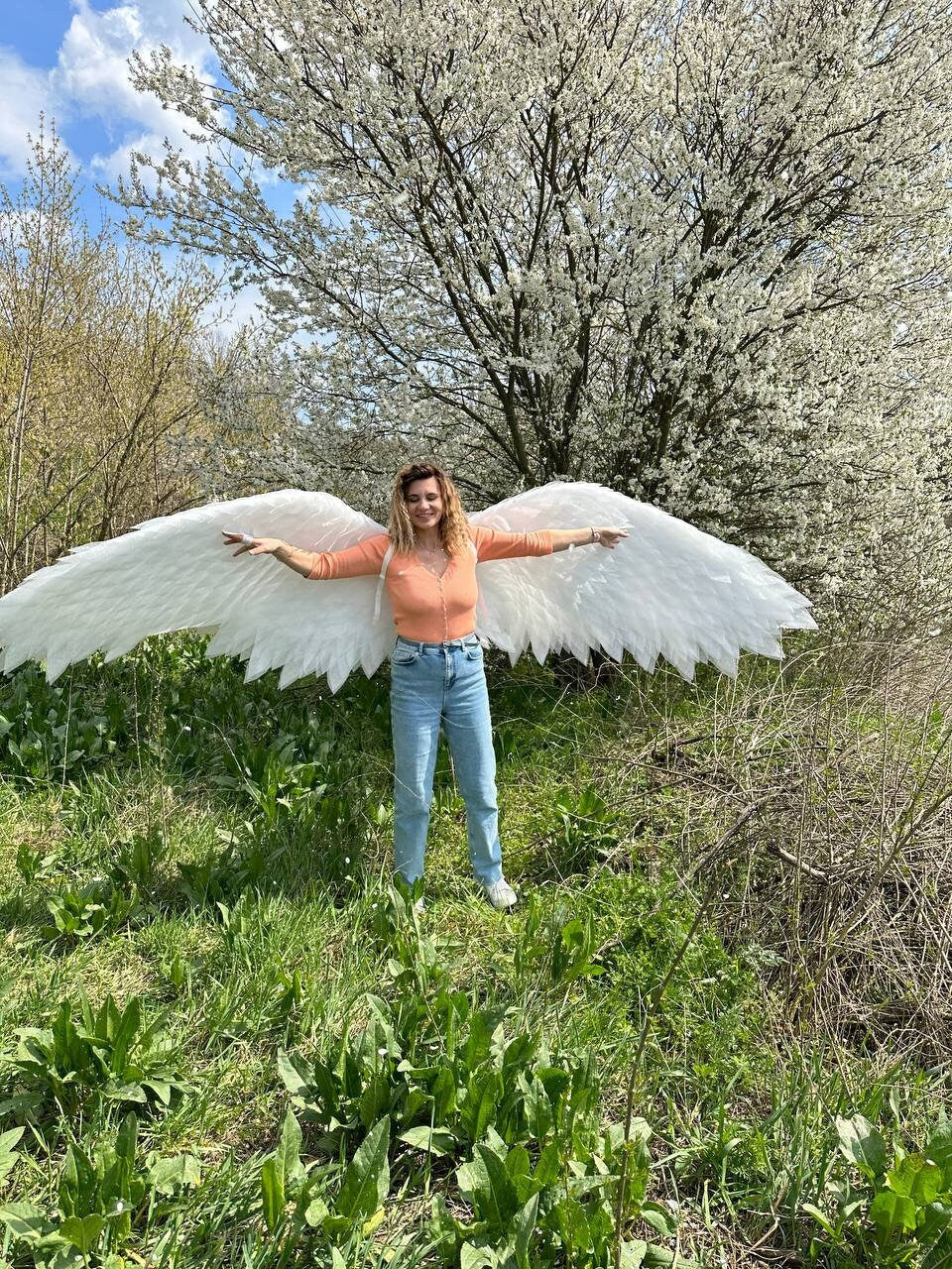 May include: A person wearing a peach colored top and blue jeans is standing in a grassy field with white angel wings spread out behind them. The person is smiling and looking at the camera. The background is a blurry image of a tree with white blossoms.