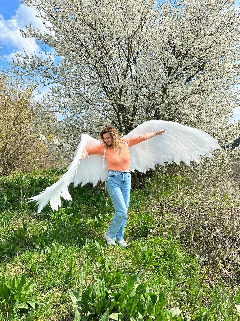 May include: A person wearing a peach colored top and blue jeans is holding large white angel wings in a field of green grass. The person is standing in front of a tree with white blossoms.