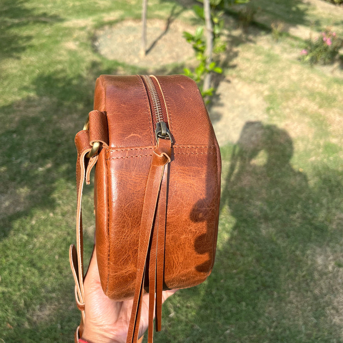 May include: A brown leather round crossbody bag with a zipper closure and a long tassel. The bag has a distressed look and is held in a hand.