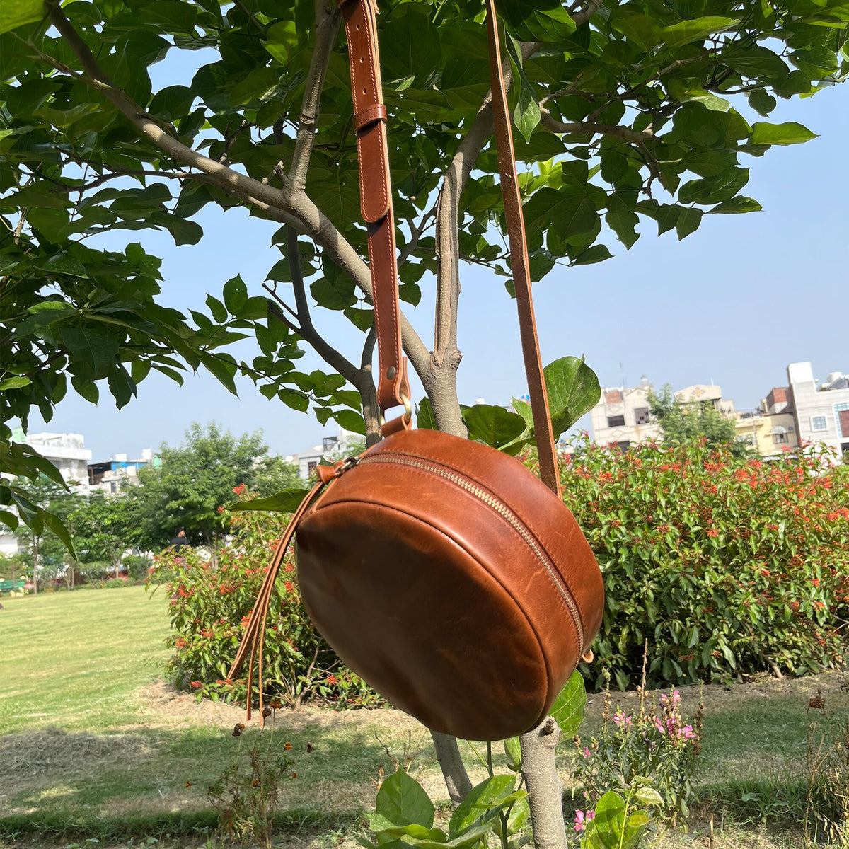 May include: A brown leather round crossbody bag with a zipper closure and a long strap. The bag is hanging from a tree branch.
