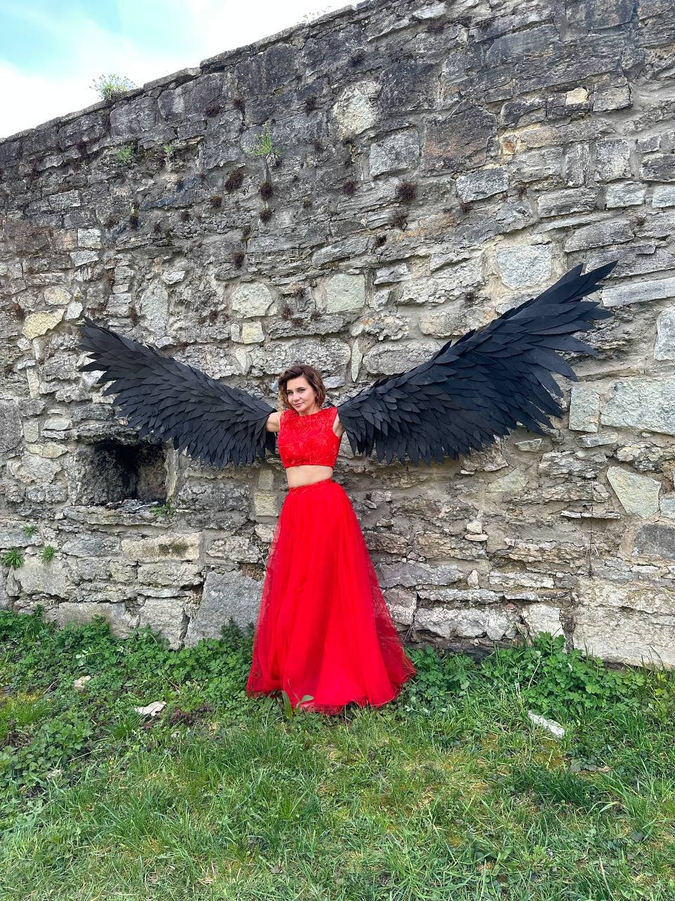 May include: A person wearing a red two-piece outfit with a black feathered angel wing costume. The person is standing in front of a stone wall with green grass in the foreground.