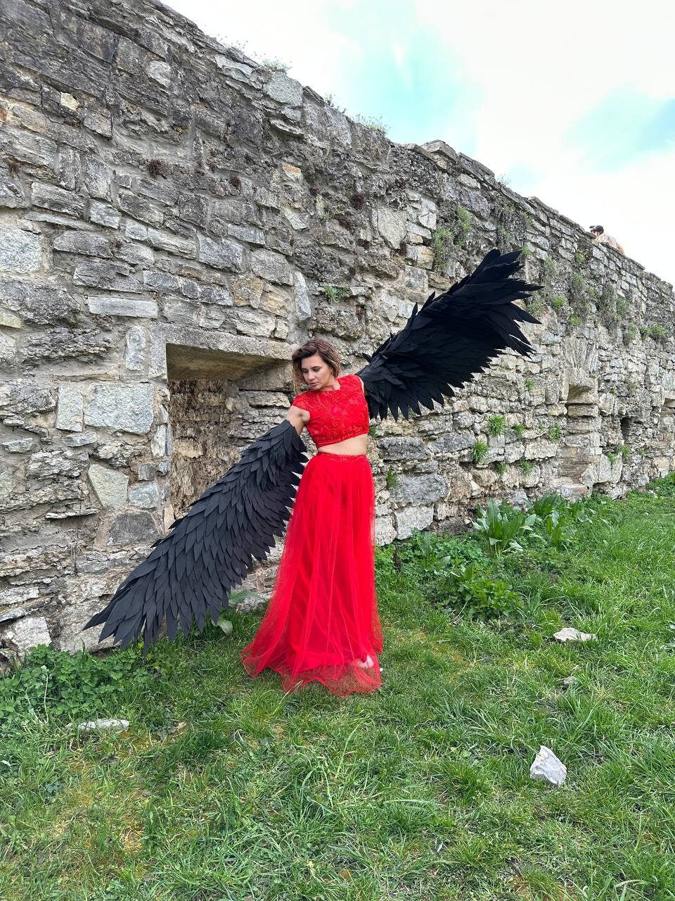 May include: A woman wearing a red crop top and a long red tulle skirt poses with large black feathered wings in front of a stone wall.