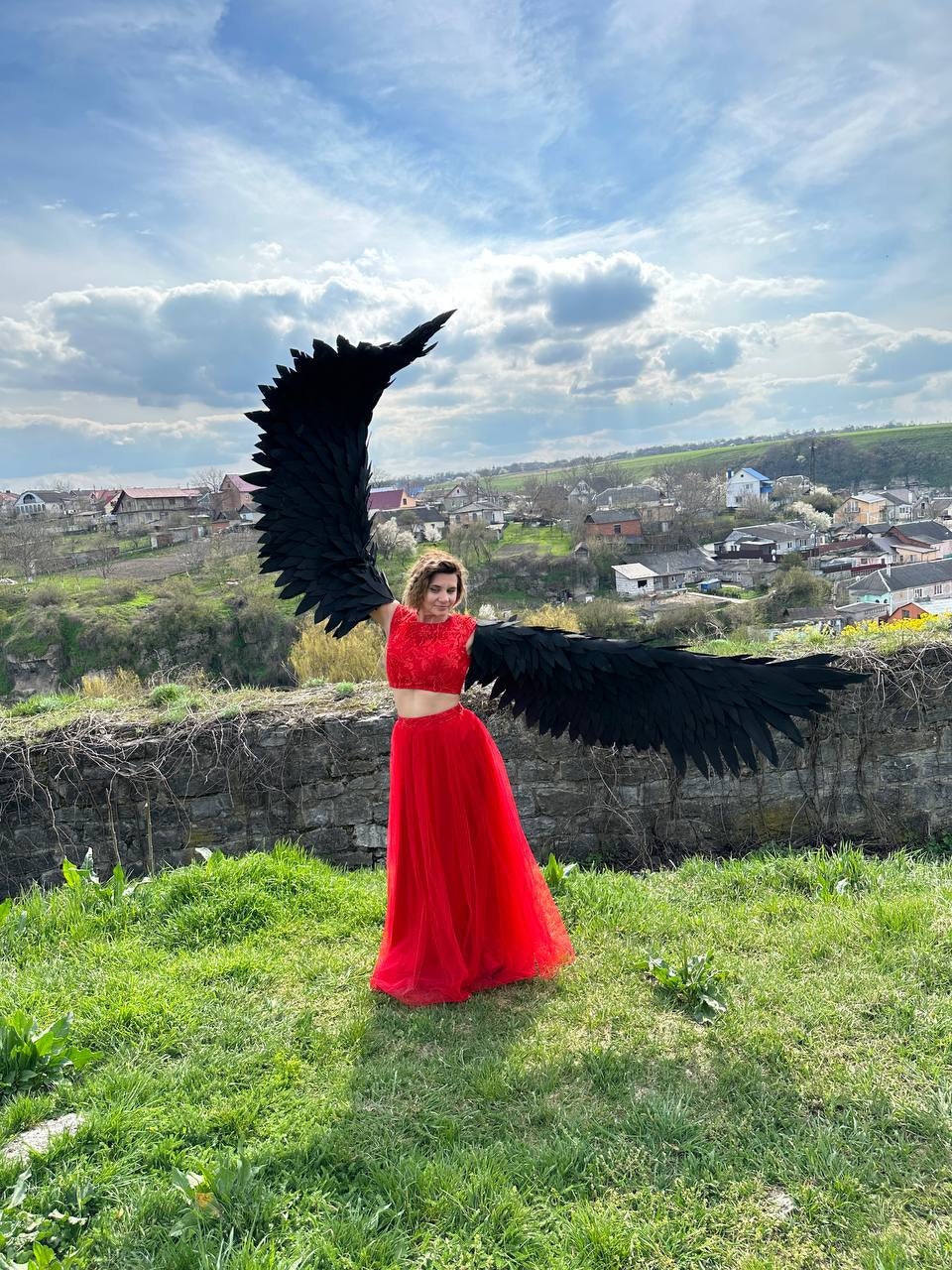 May include: A person wearing a red crop top and skirt with large black feathered wings. The person is standing in a grassy field with a village in the background.