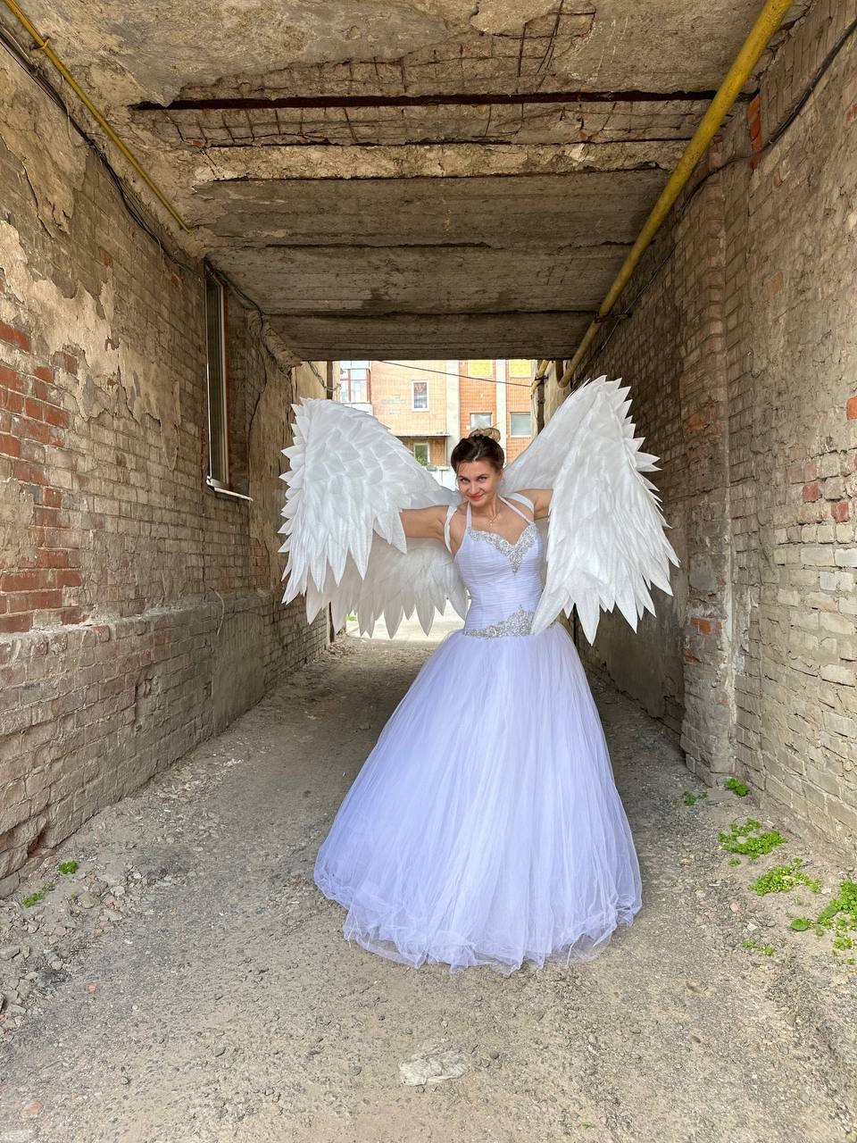 May include: A person wearing a white wedding dress and large white angel wings stands in an alleyway. The dress has a beaded bodice and a full tulle skirt. The wings are large and feathered.
