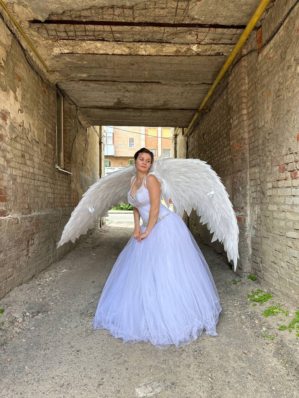 May include: A woman wearing a white wedding dress with a tulle skirt and a beaded bodice, posing with large white feathered angel wings. The photo is taken in a narrow brick alleyway, with sunlight illuminating the scene.