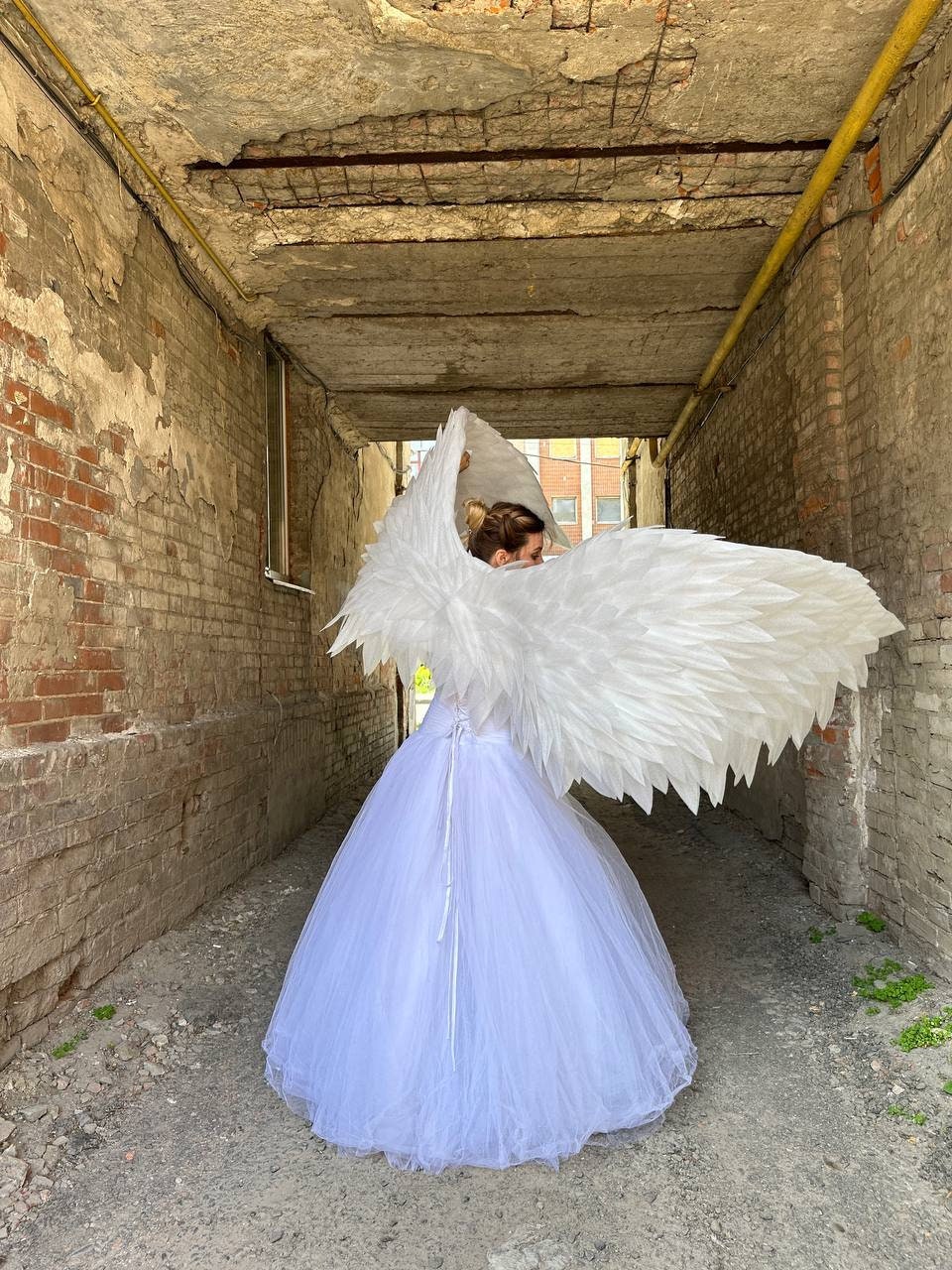 May include: A person wearing a white tulle gown and large white feathered angel wings stands in a narrow brick alleyway. The wings are spread wide, and the gown has a fitted bodice and a full skirt.