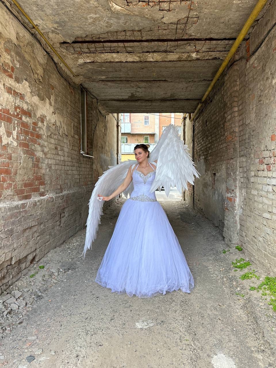 May include: A person wearing a white gown and large white feathered angel wings stands in a brick alleyway. The gown has a beaded bodice and a full, flowing skirt. The wings are spread wide, creating a dramatic effect.