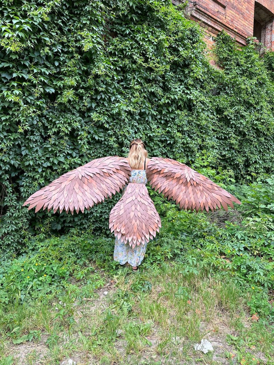 May include: A person wearing a brown leather costume with large wings. The wings are made of individual pieces of leather that are sewn together. The person is standing in front of a green ivy wall.
