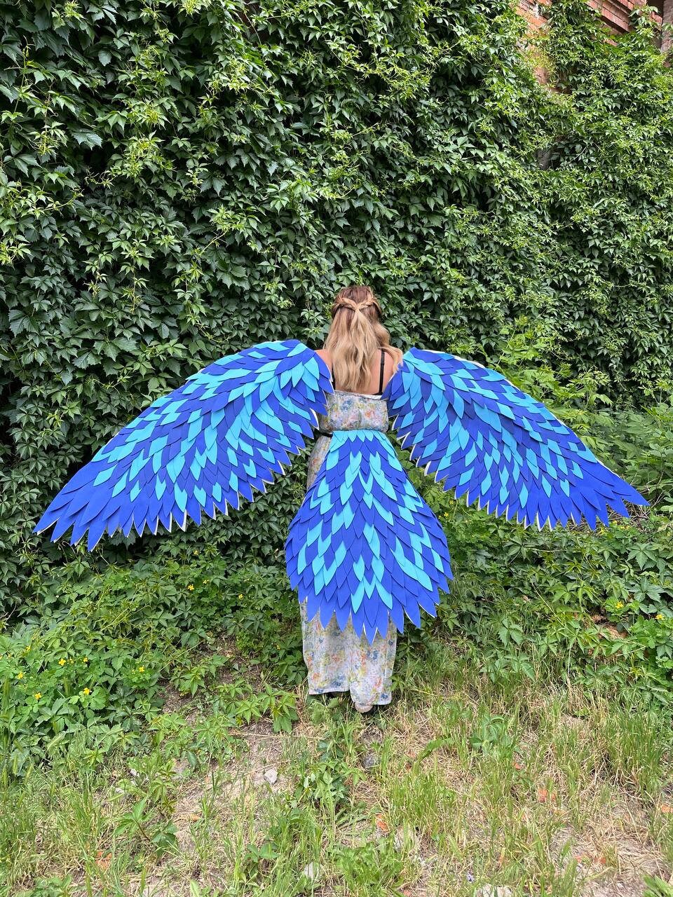 May include: A person wearing a blue and white patterned dress with large blue wings made of feathers. The wings are spread out behind the person, and they are standing in front of a green leafy wall.