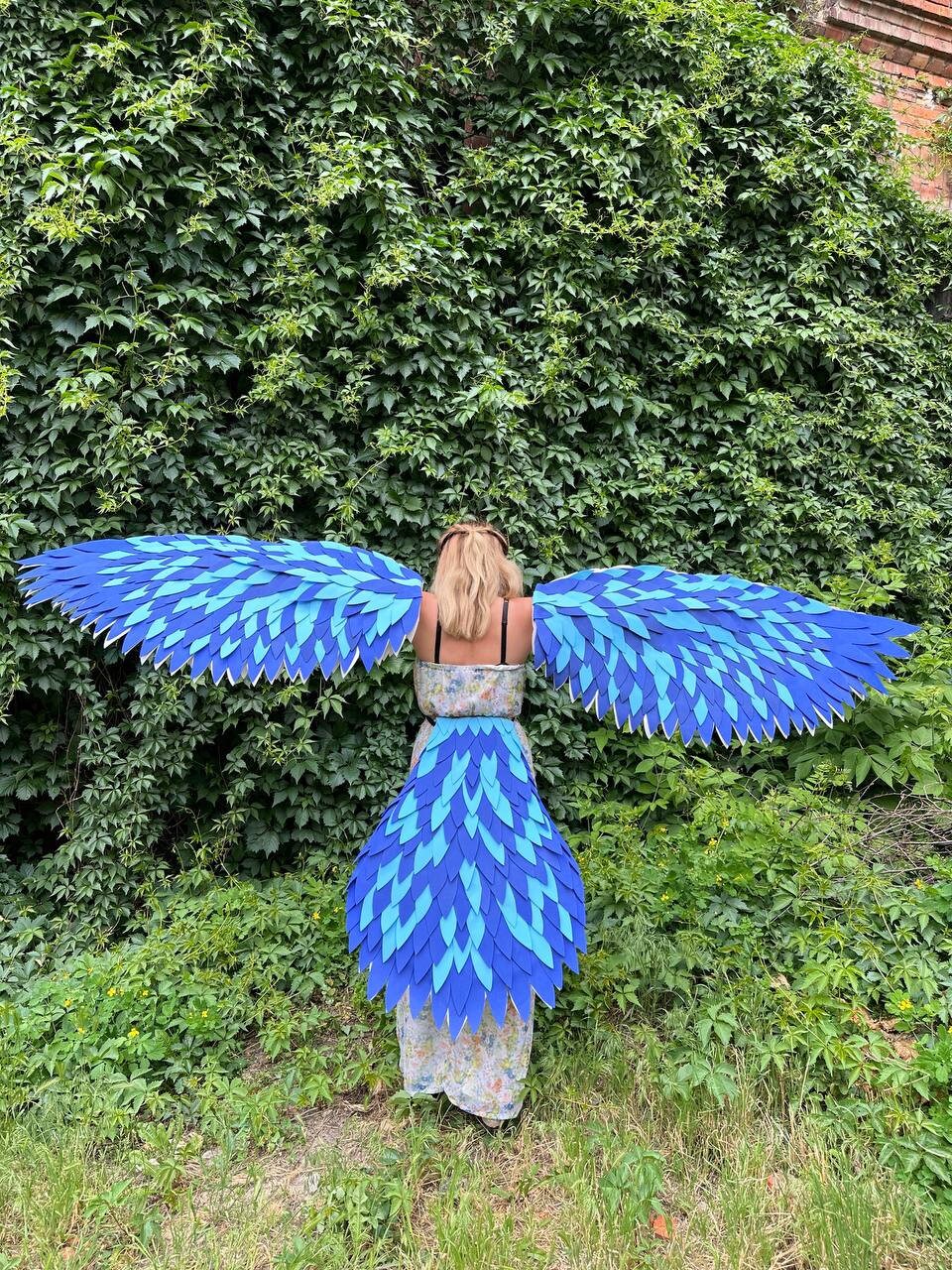 May include: A person wearing a blue and light blue feathered wings costume. The wings are large and cover the person's back. The person is standing in front of a green leafy wall.
