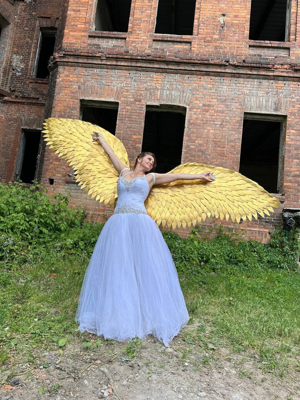 May include: A woman wearing a white tulle gown with a beaded bodice and gold angel wings. She is standing in front of a brick building.
