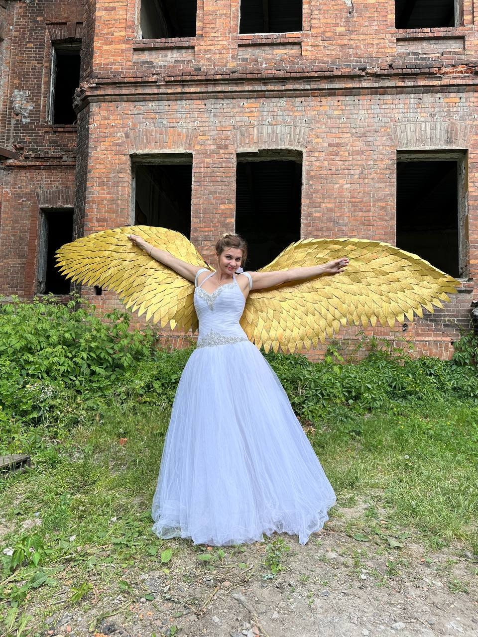 May include: A woman wearing a white wedding dress with a gold angel wing costume. The wings are made of feathers and are spread out behind her. She is standing in front of a brick building.