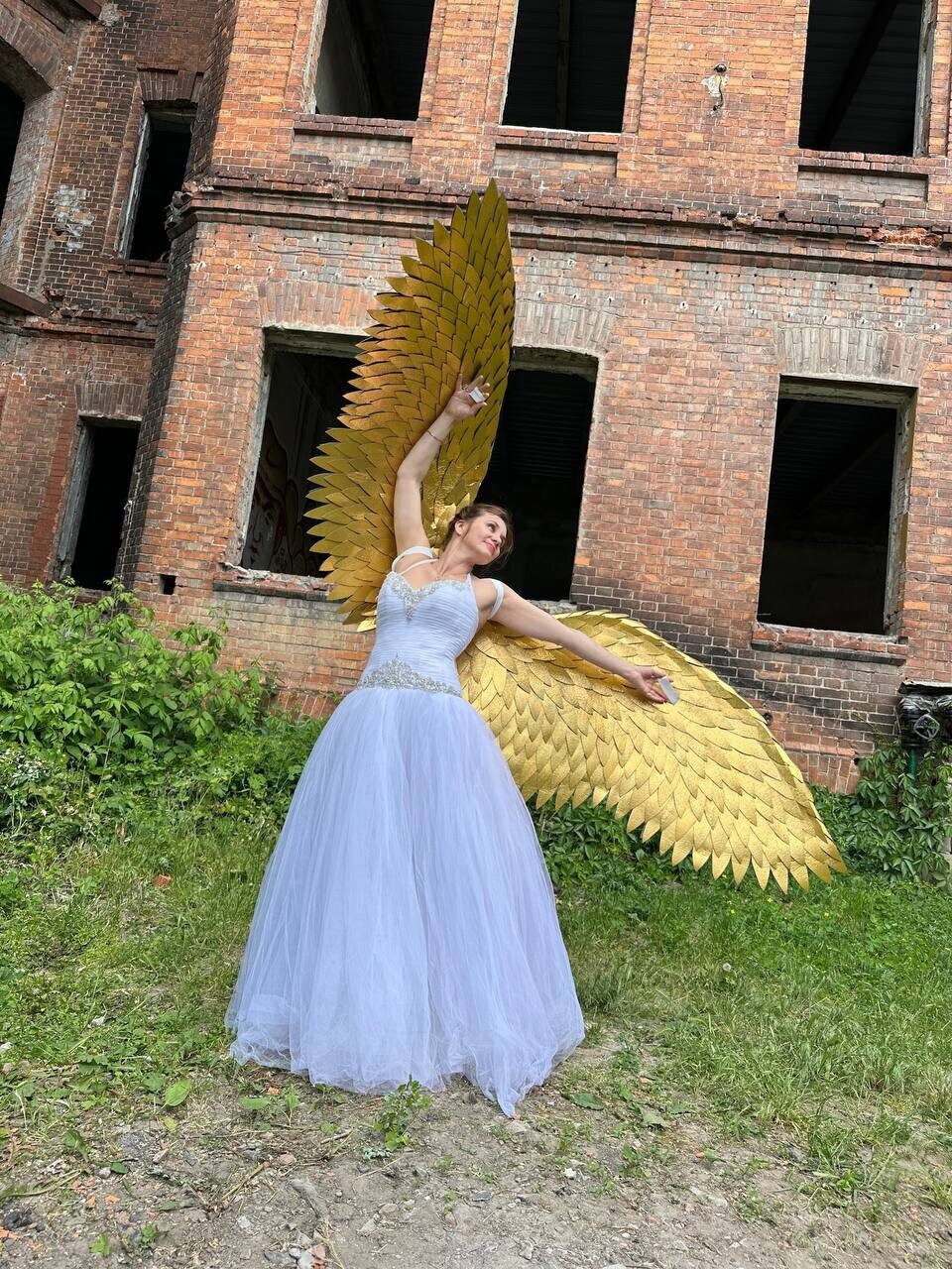 May include: A woman wearing a white tulle dress and gold angel wings stands in front of a brick building.