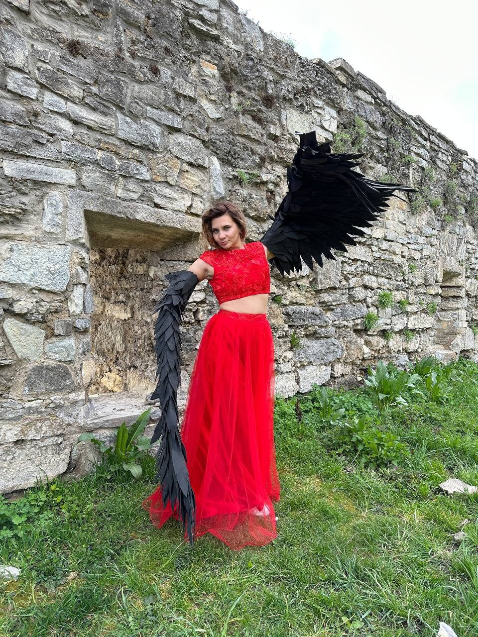 May include: A woman wearing a red crop top and a long red tulle skirt with black feathered wings and a tail. She is standing in front of a stone wall.