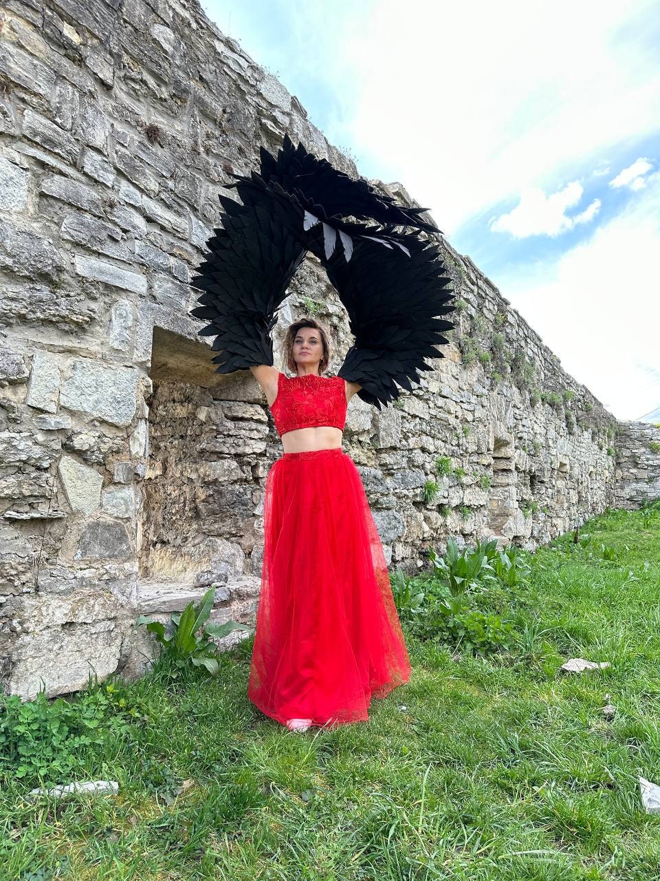 May include: A woman wearing a red crop top and a long red tulle skirt stands in front of a stone wall. She is holding large black feathered wings above her head.