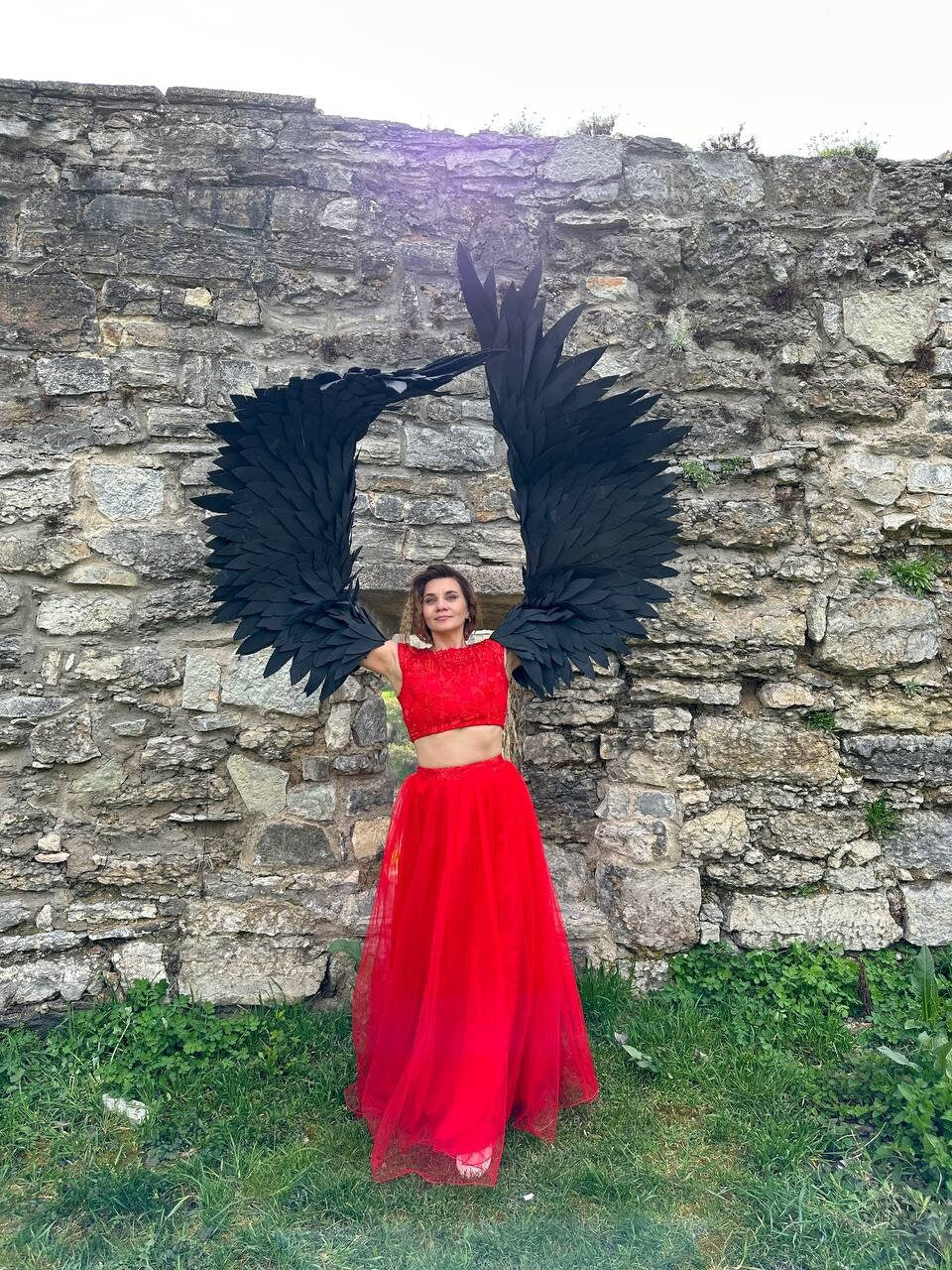 May include: A woman wearing a red crop top and a long red skirt stands in front of a stone wall. She is holding large black wings up above her head.