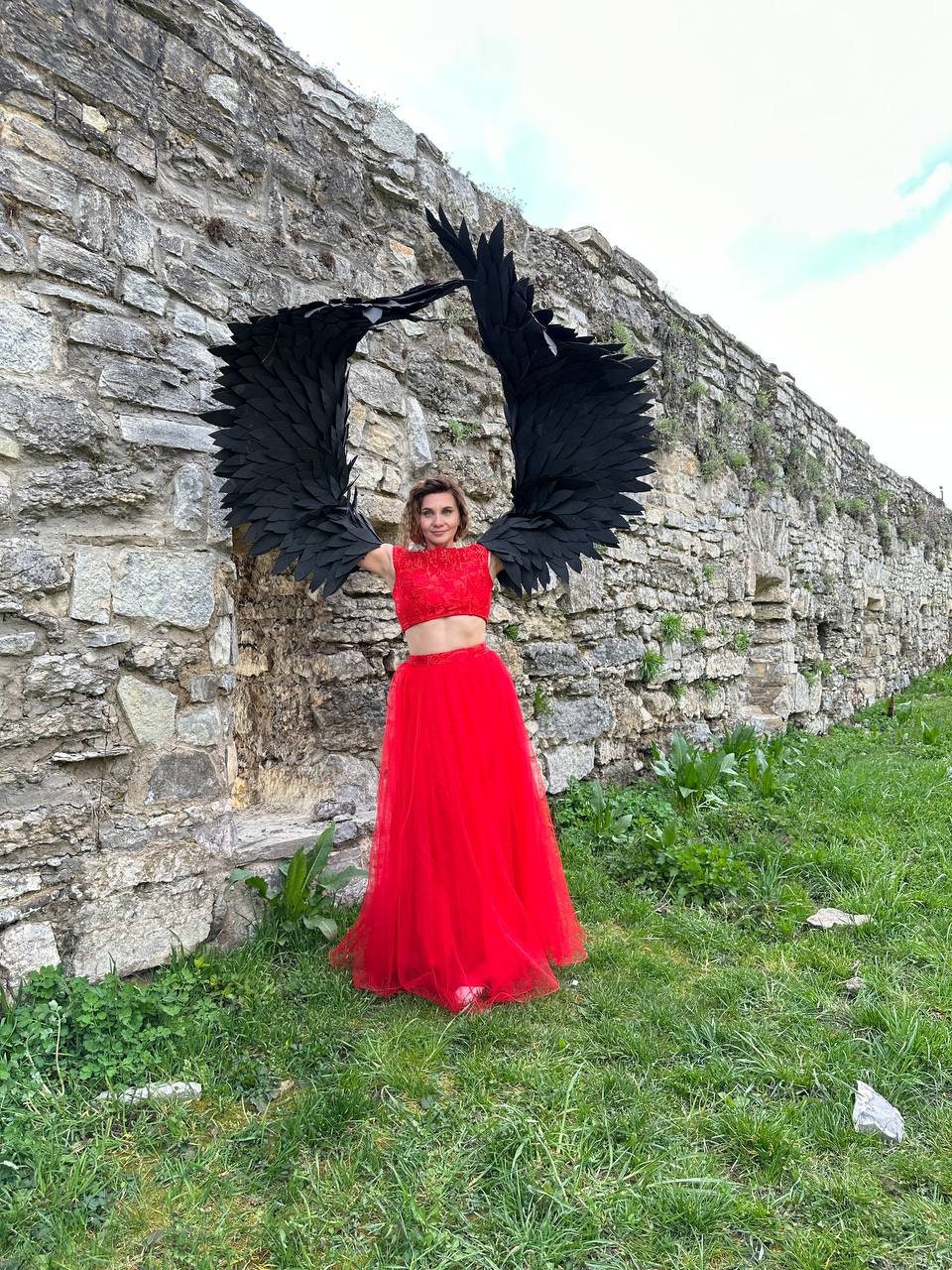 May include: A woman wearing a red crop top and a long red tulle skirt stands in front of a stone wall. She is holding large black feathered wings above her head.