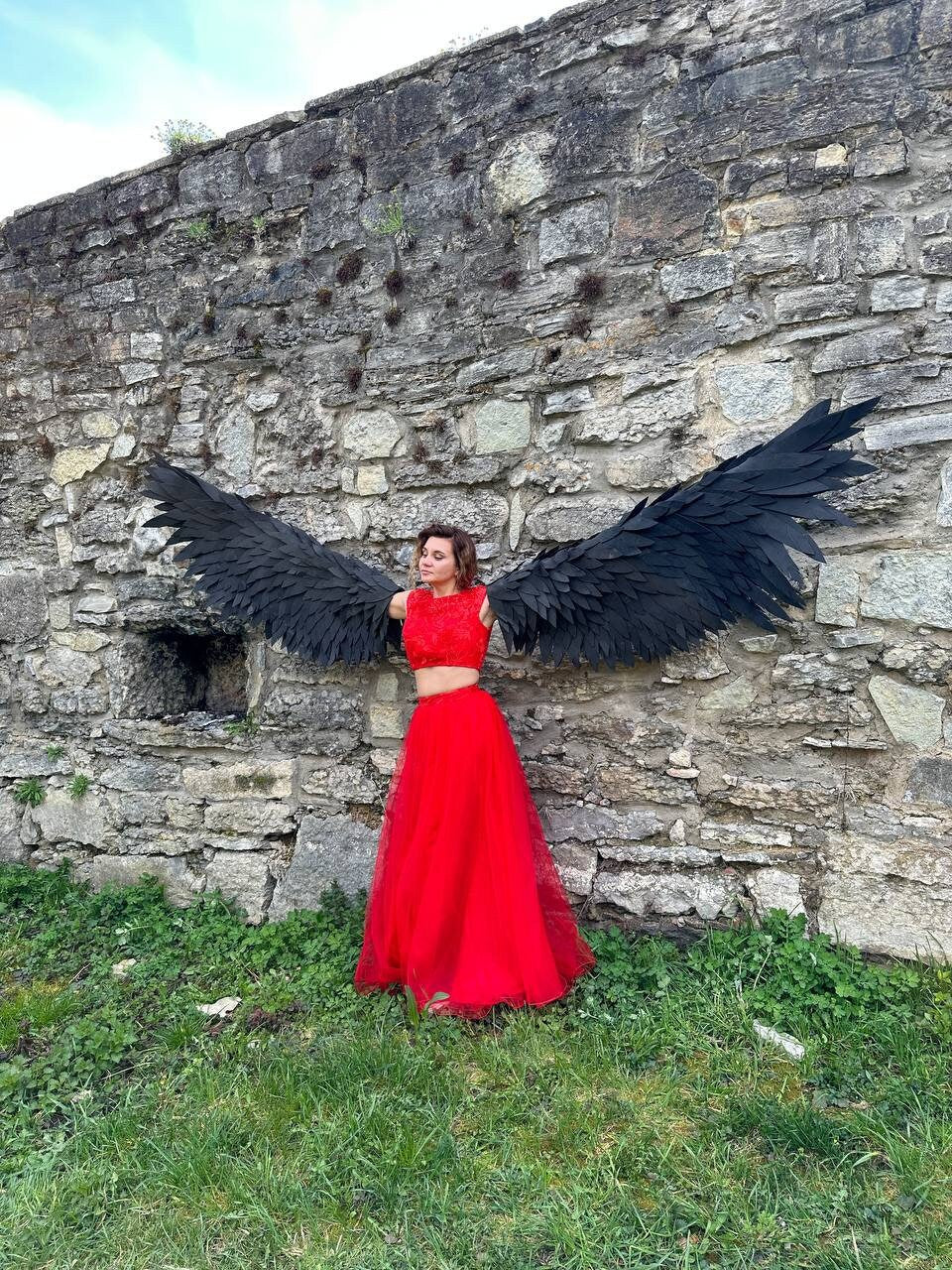 May include: A person wearing a red dress and large black wings stands in front of a stone wall.