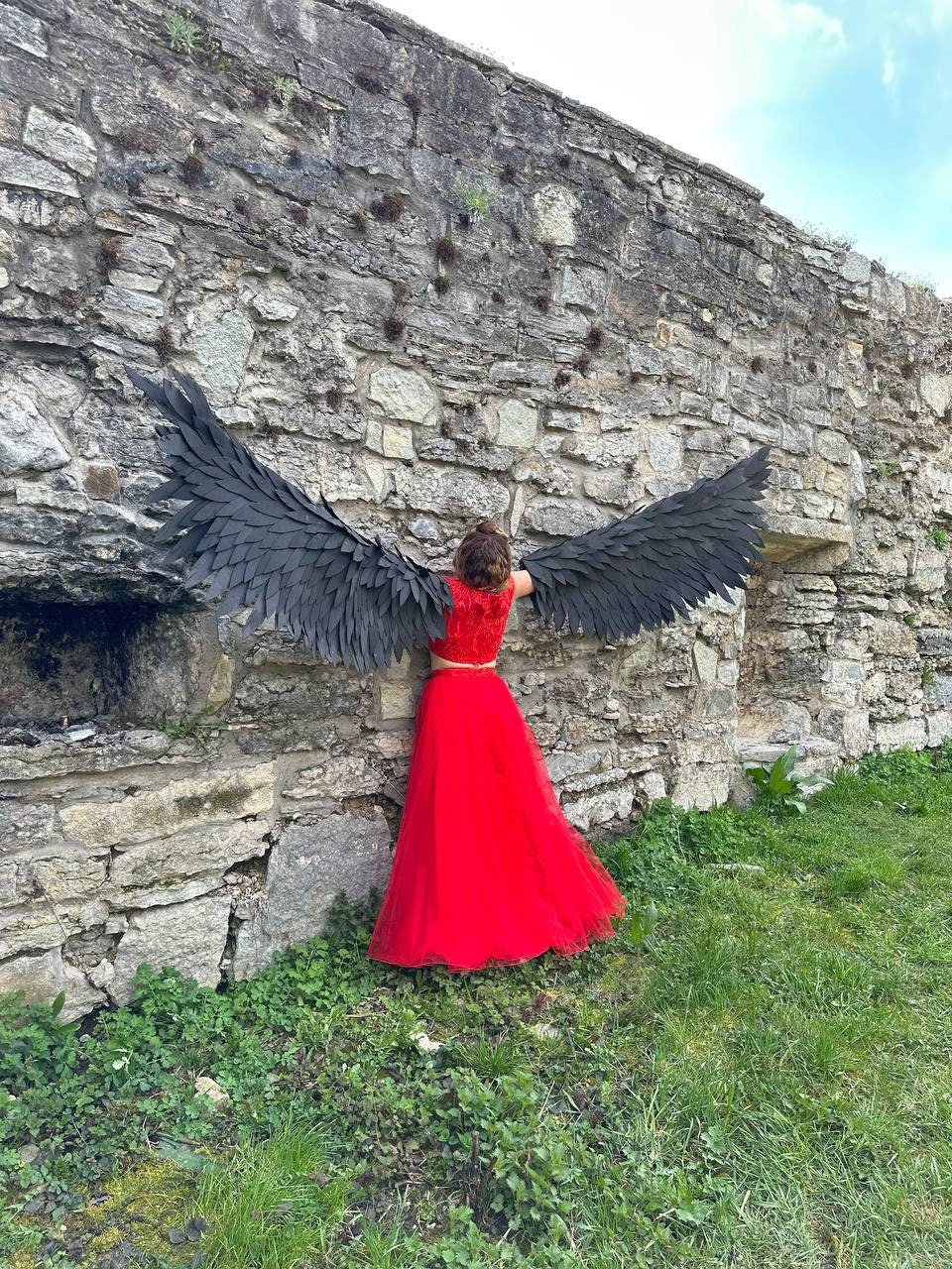 May include: A person wearing a red dress and large black angel wings stands in front of a stone wall.