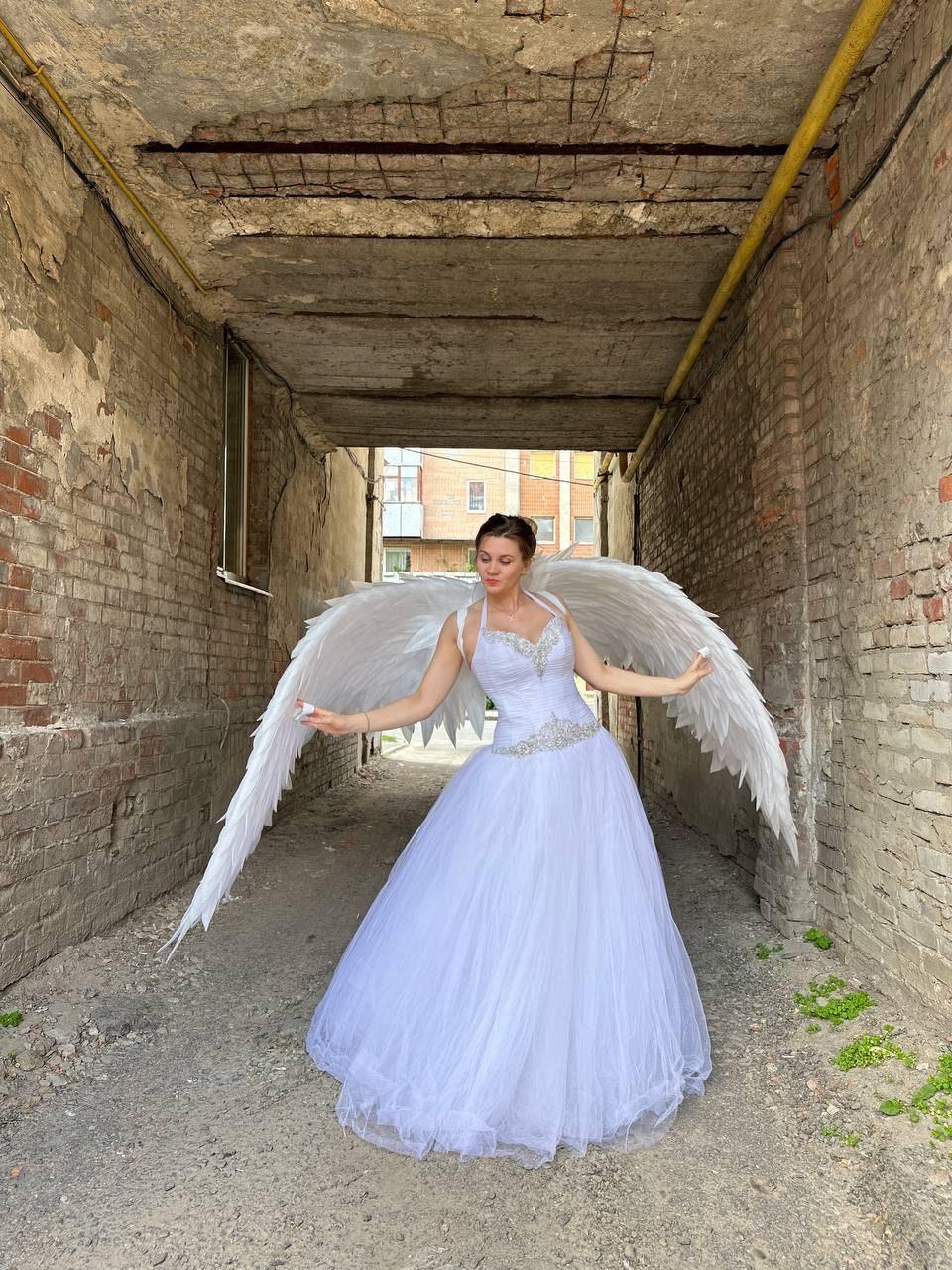 May include: A woman wearing a white wedding dress with a beaded bodice and a full tulle skirt, posing with large white angel wings. The photo is taken in a narrow, brick-lined alleyway, with the dress and wings as the focal point.