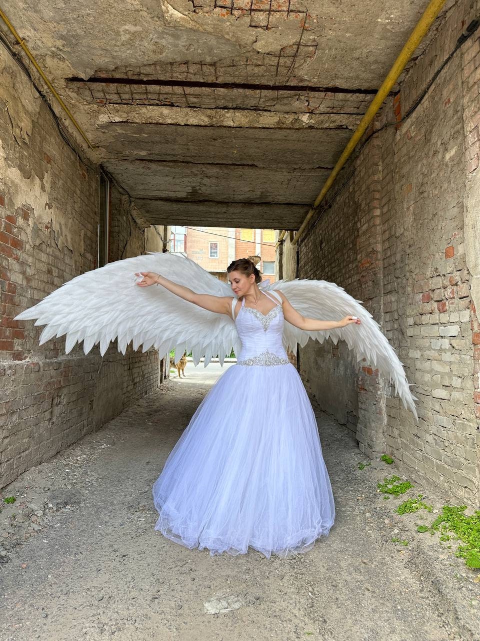 May include: A woman wearing a white, floor-length gown with a beaded bodice and a full tulle skirt. She is wearing large, white feathered angel wings. The photo is taken in a narrow alleyway with brick walls.