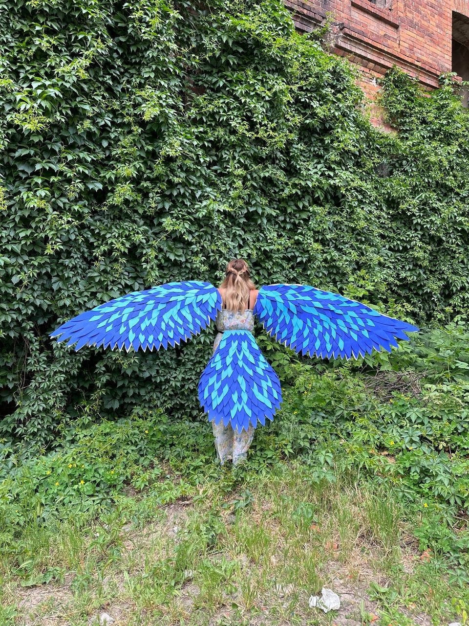 May include: A person wearing a blue and green costume with large wings. The wings are made of feathers and are spread out behind the person. The person is standing in front of a green wall.