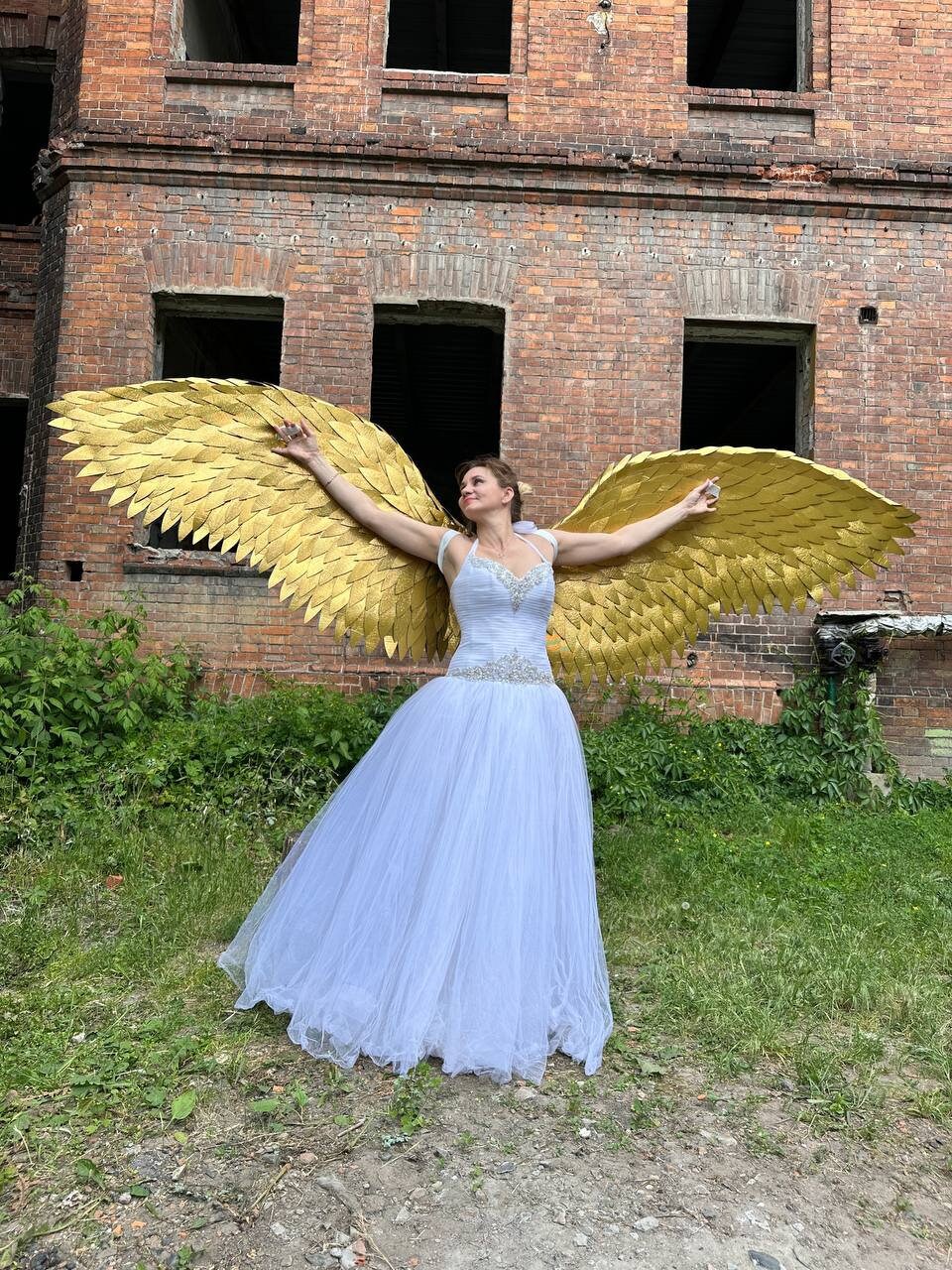 May include: A woman wearing a white wedding dress with a gold angel wing costume. The wings are large and feathery, and the woman is standing in front of a brick building.