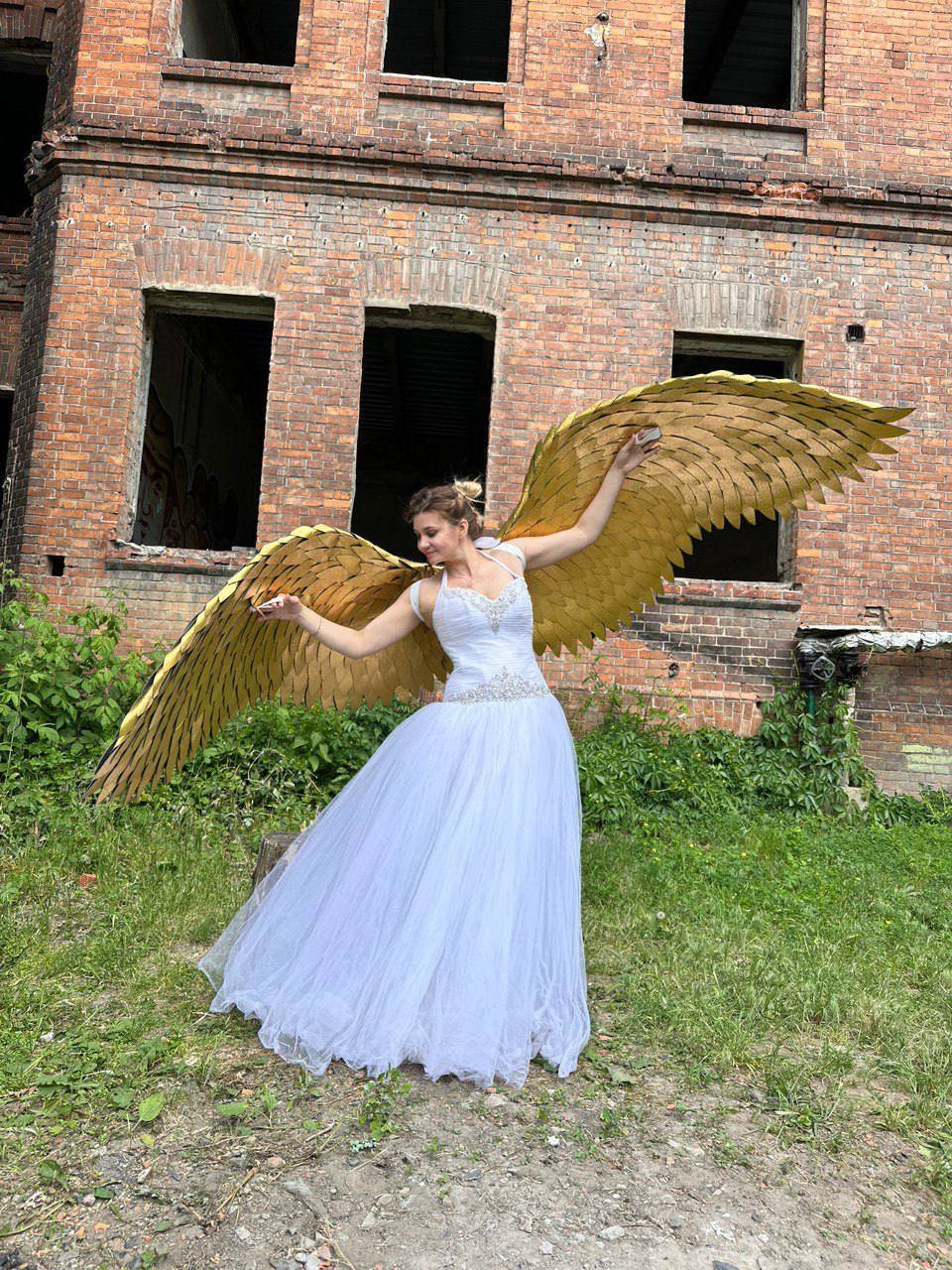 May include: A woman wearing a white dress and large gold angel wings, standing in front of a brick building.